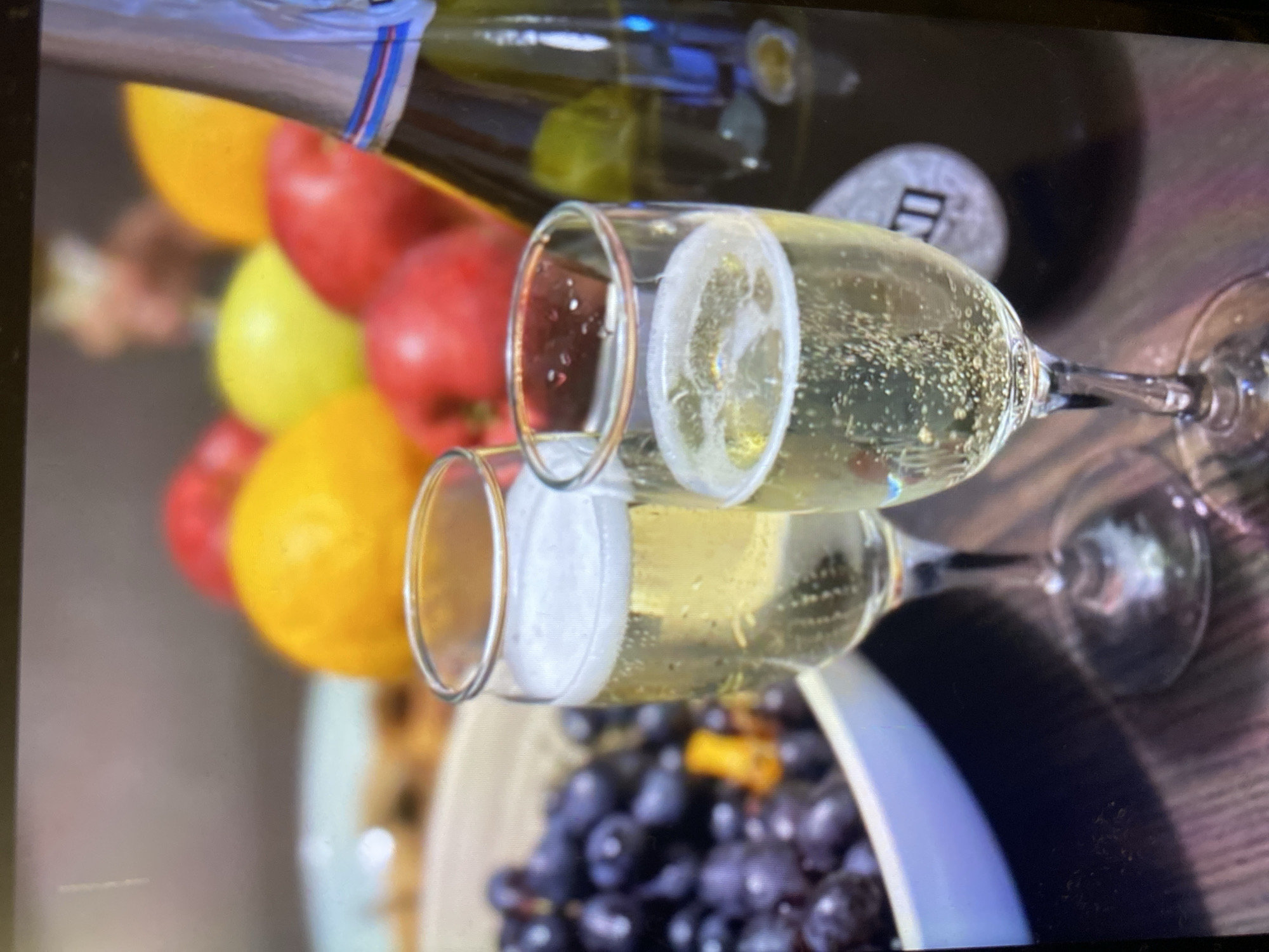 Two champagne flutes filled with a bubbly, pale yellow liquid are positioned in the foreground. Behind them, a bottle of champagne with a silver foil cap stands upright. A bowl of dark purple grapes is to the left, and various fruits, including apples and possibly a lemon or orange, are visible in the background. The scene appears to be indoors, possibly on a table with a wooden surface, and the lighting suggests it might be evening. There is no identifiable information to indicate a specific location in Kamianske, Ukraine.