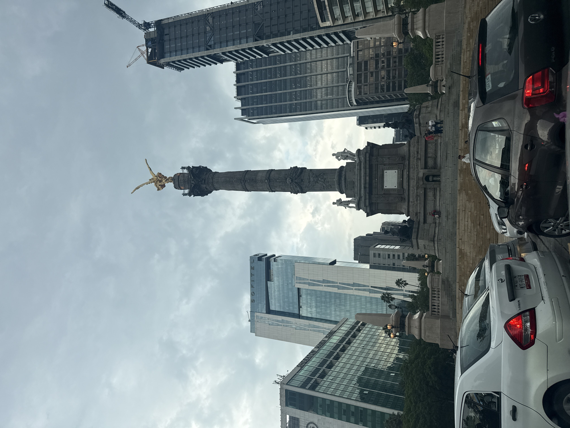 The image captures the Ángel de la Independencia (Angel of Independence) monument in Mexico City, Mexico, positioned centrally within a roundabout amidst dense urban development. The monument itself is characterized by a tall column topped with a golden statue of the Winged Victory. Multiple buildings flank the monument, including modern skyscrapers under construction and established structures like the Sheraton Maria Isabel Hotel. Traffic, including a white Nissan Tsuru, surrounds the monument on the lower right side of the frame. The sky is overcast with dense cloud cover. There are people near the base of the monument.