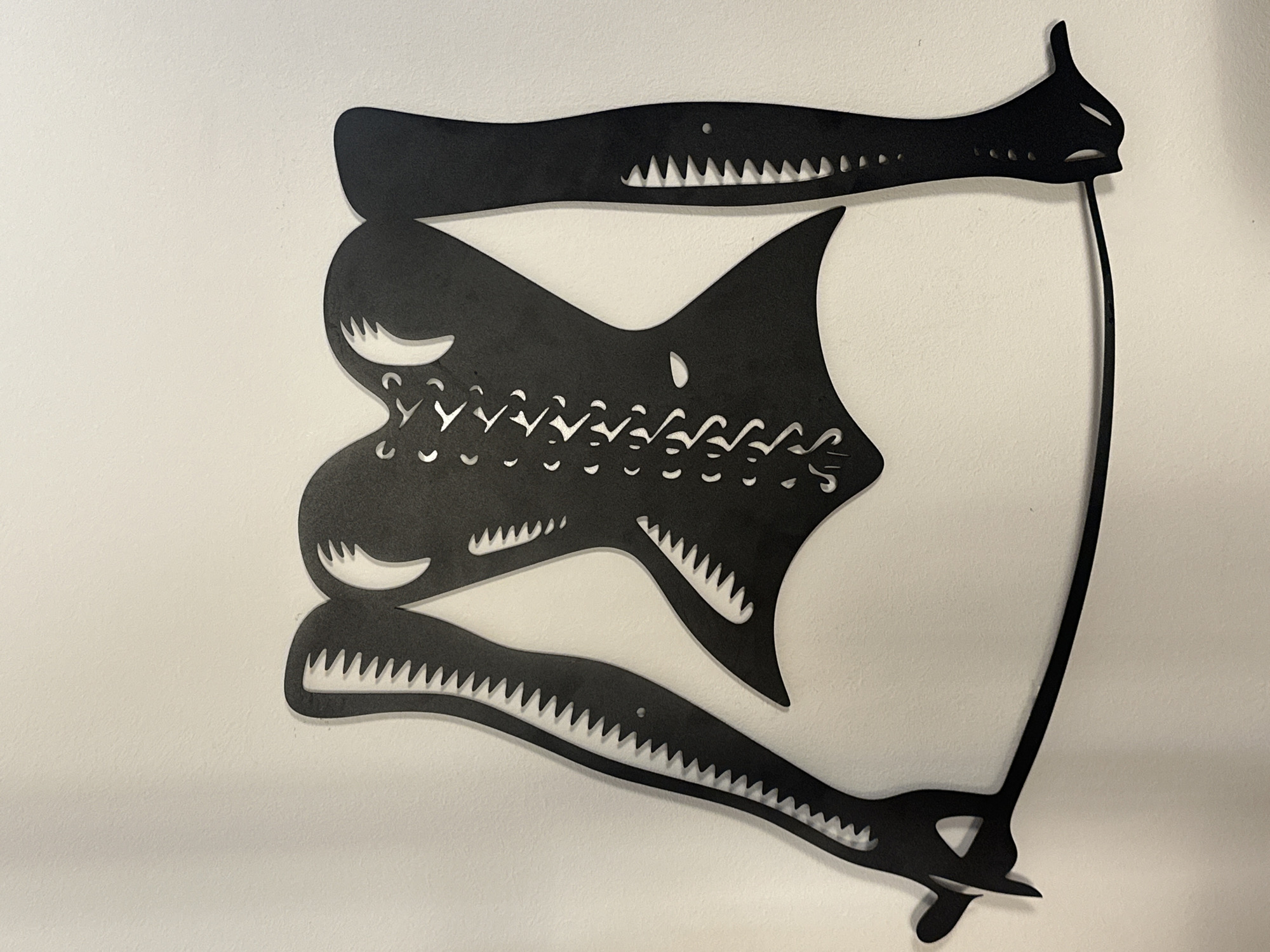 A black silhouette of a female torso and arms, styled as a corset with lace-up details, is mounted on a white wall. The silhouette is abstract and stylized, with sharp, angular edges and cut-out details. The arms extend outwards, with one hand seemingly holding a thin, curved object that runs along the bottom of the frame. The object appears to be a whip or a similar implement. The wall is a plain white surface, providing a neutral background that emphasizes the black silhouette. The lighting is even, casting minimal shadows. The image is a close-up view, with the full extent of the silhouette filling most of the frame. There are no discernible features of Budva, Montenegro, visible in the image.