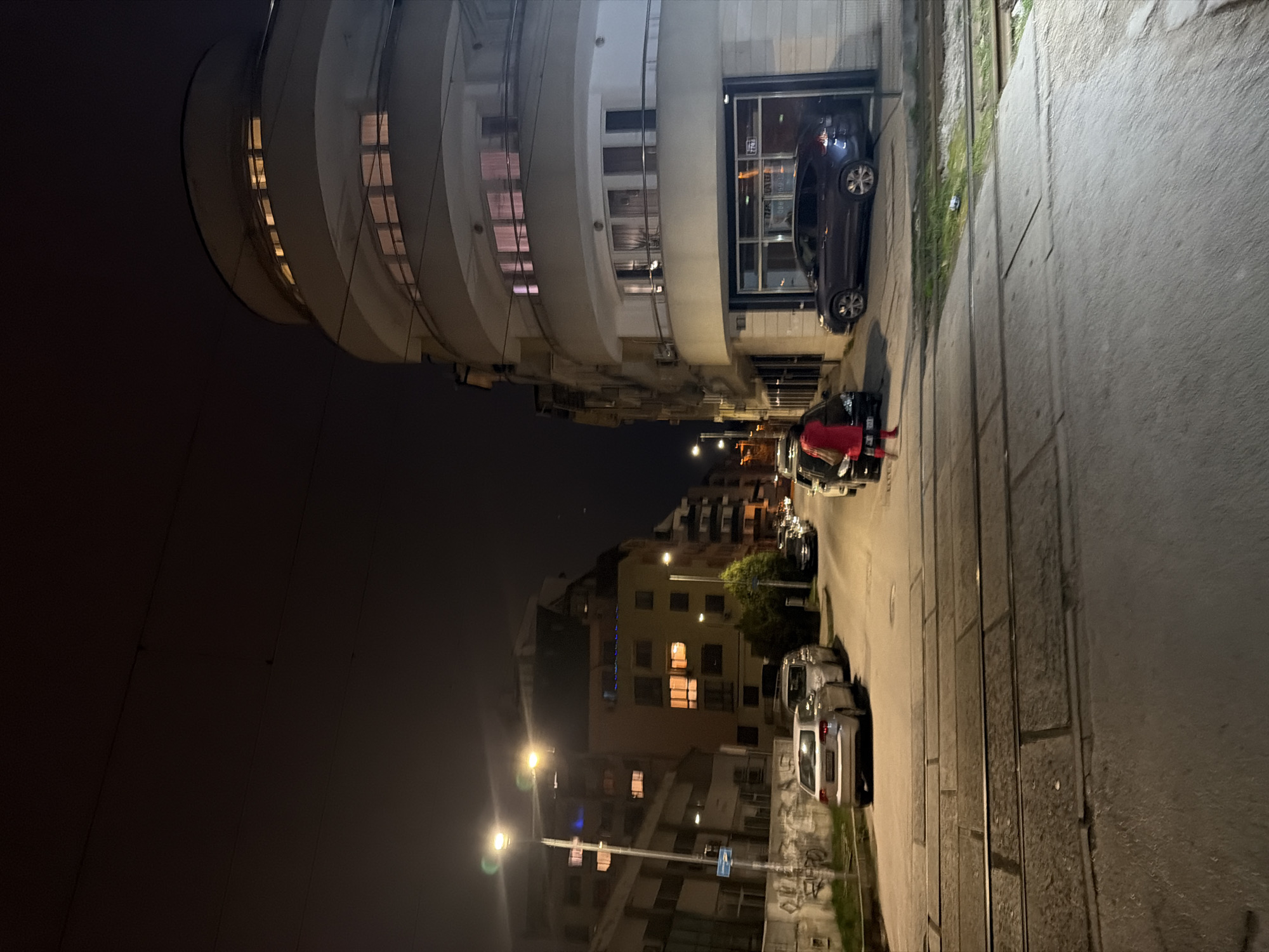 Here is a description of the image:
The image depicts a street scene at night in Sofia, Bulgaria. The architectural landmark is the "Raya" office building at 136 Tsar Boris III Boulevard. The photo is taken from the street looking towards the Raya building. Cars are parked along the street. A person in a red jacket is standing near a vehicle. Street lights illuminate the scene. Overhead tram lines run along the street. The background features other residential and commercial buildings, creating an urban environment.