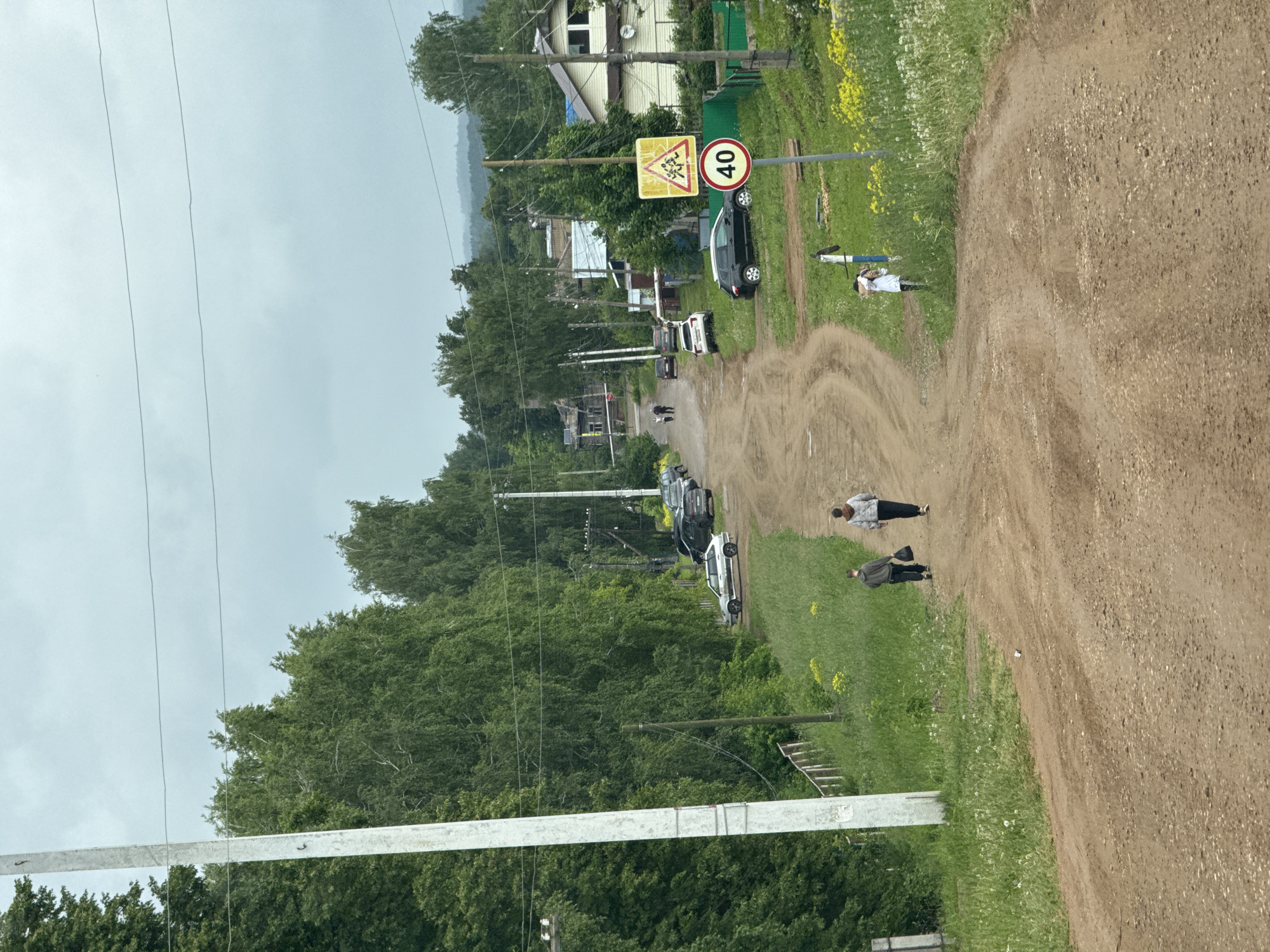 The image captures a rural or semi-rural scene in Фоки, Russia, under an overcast sky during daytime. A wide, unpaved dirt road, marked with vehicle tracks, dominates the foreground and extends into the distance, suggesting a village street. On the left side of the road, a grassy verge with patches of yellow wildflowers separates the thoroughfare from a line of parked cars and dense green trees. Behind the trees, several residential buildings are partially visible.
Overhead, multiple power lines crisscross the pale sky, supported by wooden poles. A metal horizontal bar is also present in the lower left foreground. A prominent pole near the road features two signs: a yellow triangular warning sign depicting two children, indicating a "Children Crossing" area, and a red-bordered circular speed limit sign displaying "40", signifying a 40 km/h maximum speed.
Several people are visible in the scene. Two adults are walking along the dirt path; one wears a light-colored top and dark pants, while the other, in darker attire, carries a bag. Further ahead on the grassy area, a person in a white shirt stands next to a blue object, possibly a bicycle or scooter. Multiple cars, including a black SUV and a white car, are parked along the left shoulder of the road. The scene conveys a sense of everyday activity in a quiet, residential village setting.