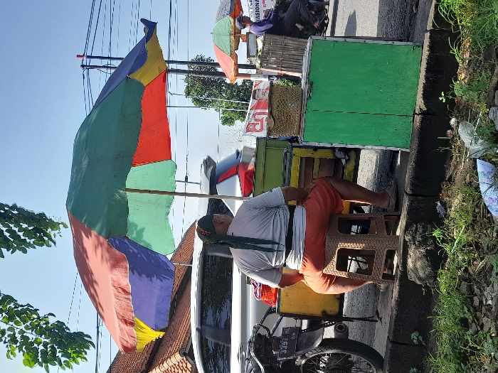 An elderly man sits on a brown plastic stool next to his mobile food stall. He wears a white shirt, orange shorts, and a green head covering. His stall is attached to a motorcycle with license plate 3292 HL and is covered by a colorful umbrella. A second stall with an umbrella sits behind his. Both are parked along a curb lined with weeds and debris. The sky is blue and clear.