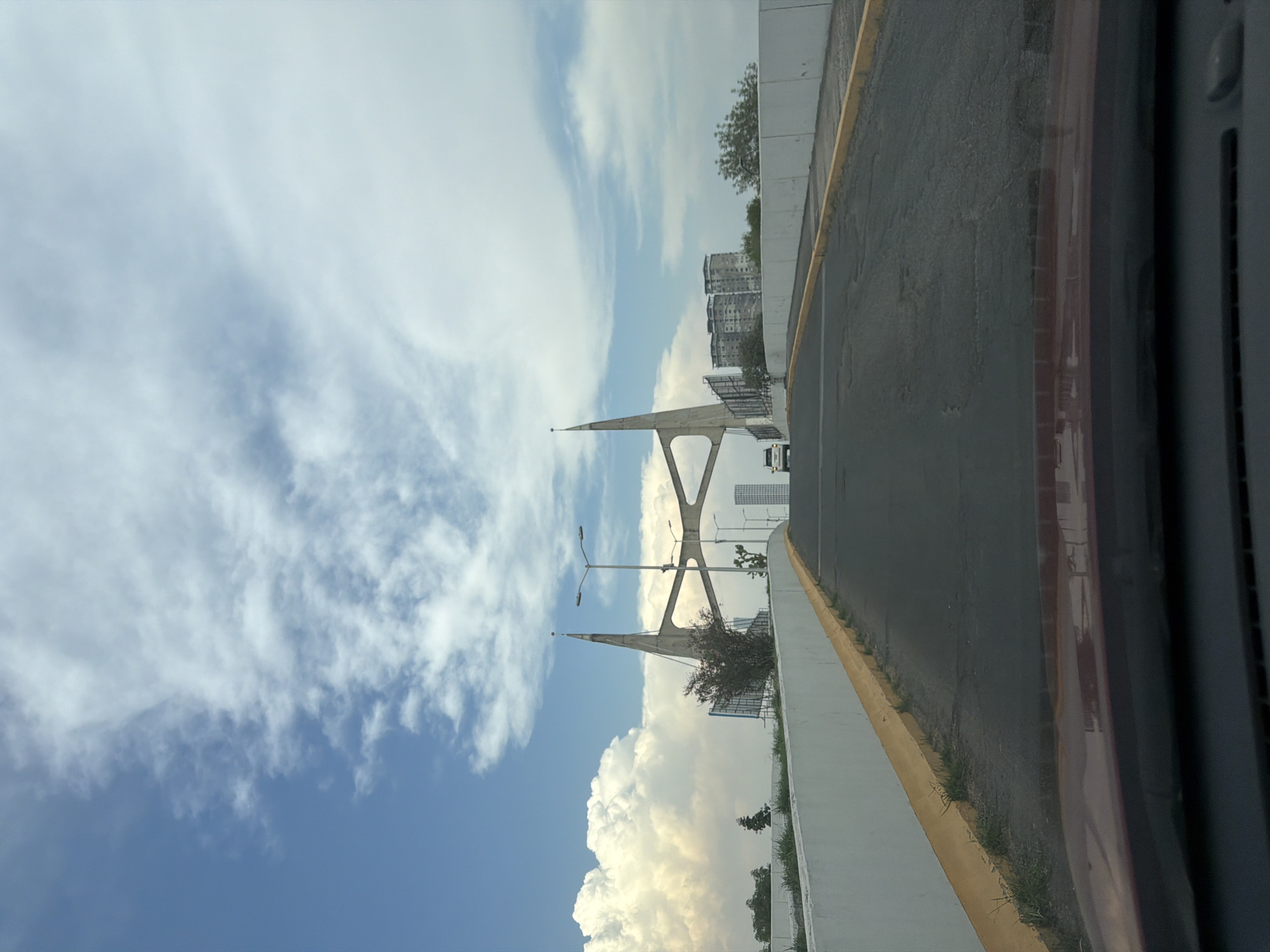 The image shows a view from a vehicle on a road in San Bernardino Tlaxcalancingo, Mexico. Visible on the left is a section of the cable-stayed bridge called the Viaducto Elevado. The sky above is filled with cumulus clouds and clear blue areas. The road is asphalt, with a white concrete barrier on the left and a yellow curb separating it from a shoulder. The vehicle's window frame is visible on the right. In the background, buildings and more of the bridge's structure are visible.