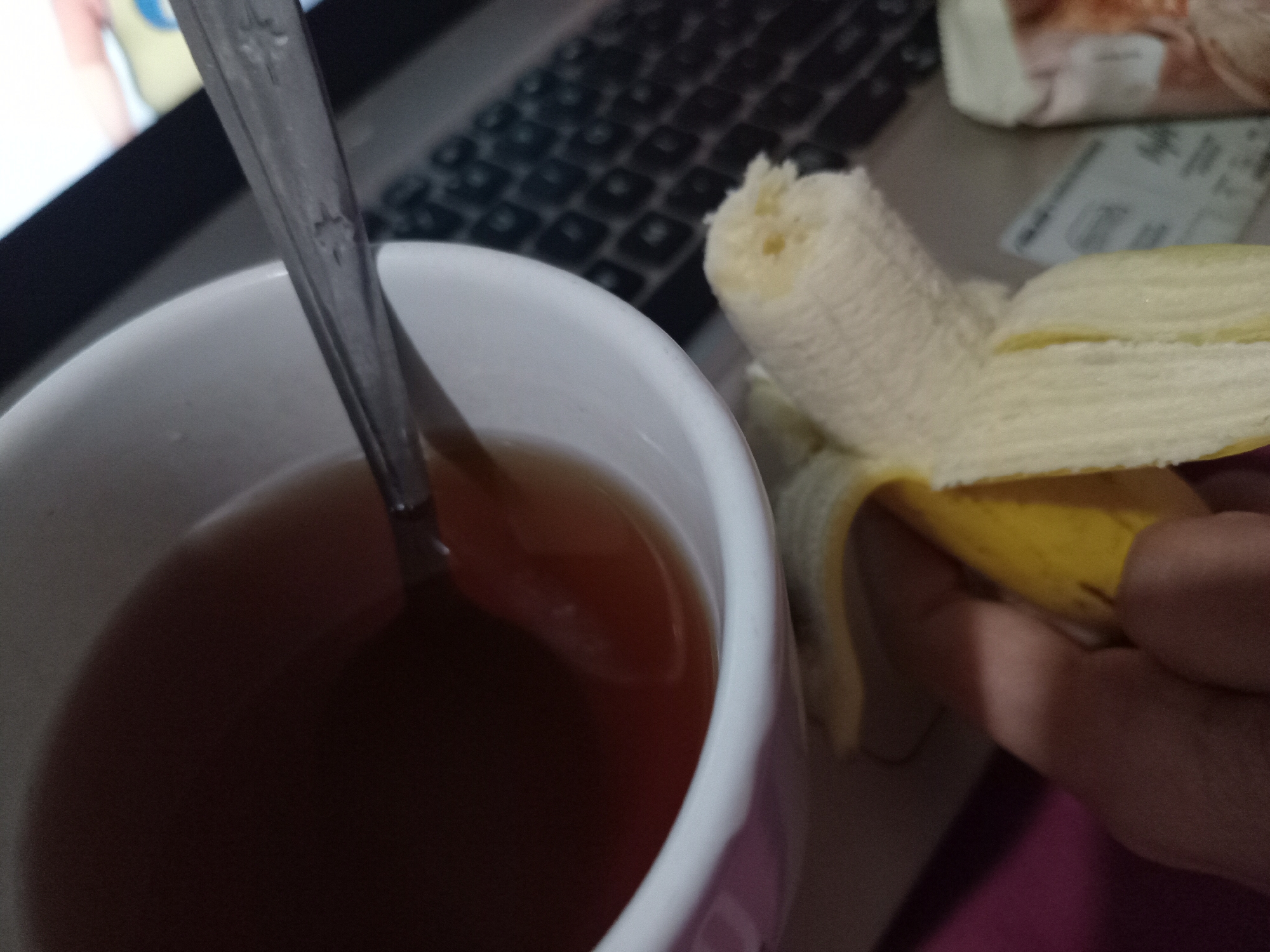 In Claket, Indonesia, a close-up shot focuses on a white mug filled with a dark beverage, likely tea, with a silver spoon submerged within. To the right of the mug, a partially peeled banana is held by a hand, its yellow peel folded back to reveal the white fruit. The background shows a blurred laptop keyboard, suggesting a workspace or study area. The lighting is dim, casting shadows and creating a warm, intimate atmosphere.