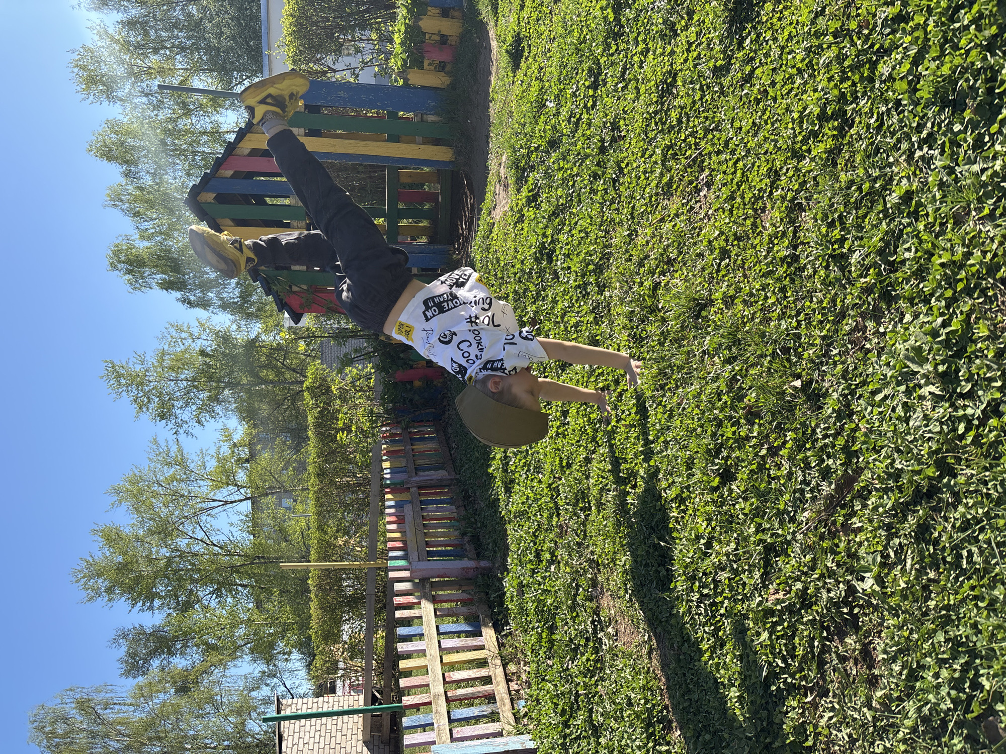 A boy is performing a handstand on a sunny day in what appears to be a grassy area. He is wearing a white t-shirt with graphic text and designs, dark pants, yellow shoes, and a green cap. His body is inverted, with his legs pointing upwards and his hands touching the ground. In the background, there's a colorful wooden structure resembling a climbing frame or fence. There are trees and a clear blue sky visible in the background as well. The location is outdoors in Chaikovsky, Russia.