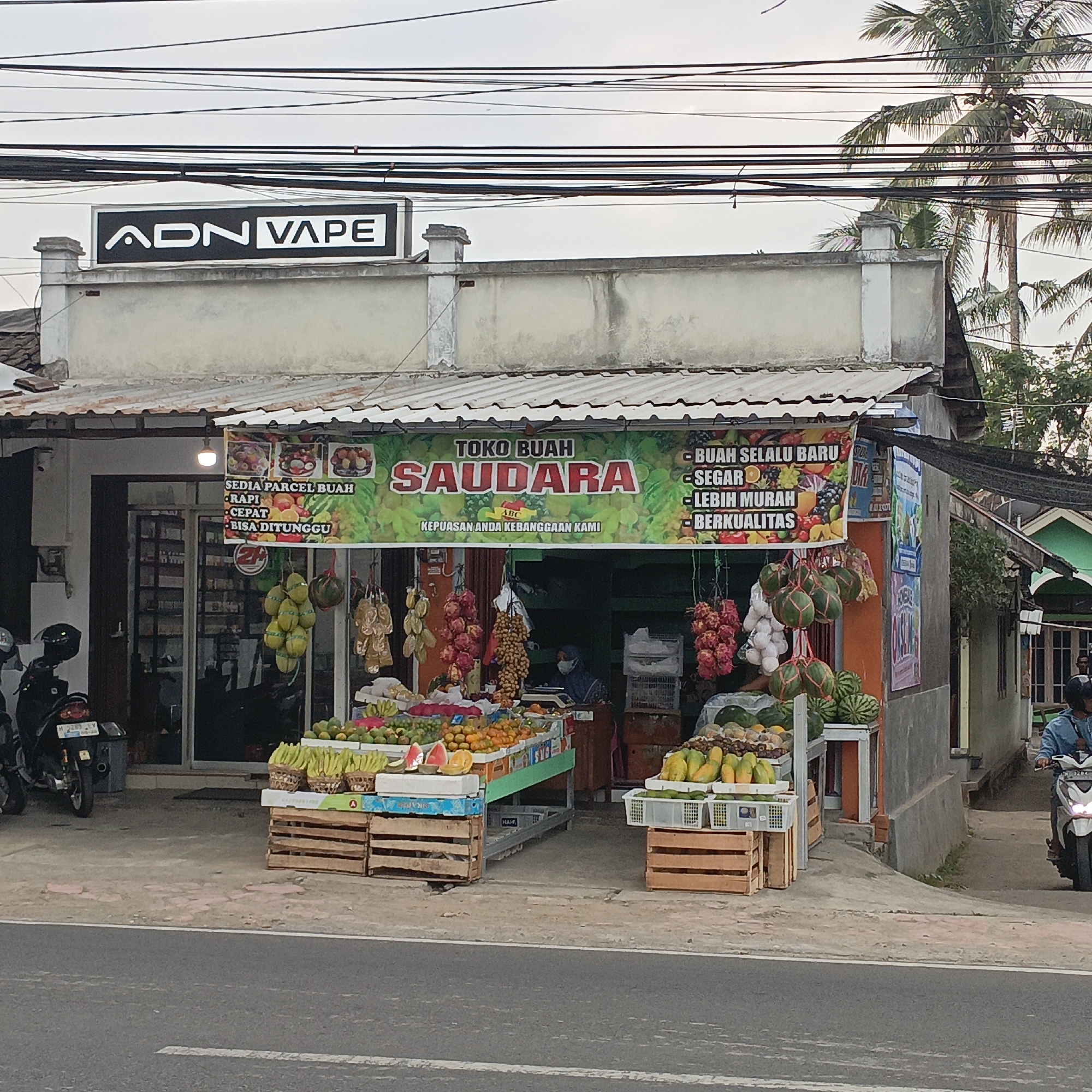 A fruit stall named "Toko Buah Saudara" is displayed on the side of a road in Salatiga, Indonesia. The stall features a variety of fresh fruits such as watermelons, papayas, bananas, oranges, and dragon fruits, arranged in wooden crates and baskets. Behind the stall, the entrance to a shop is visible, with shelves stocked with goods. A banner above the fruit stall advertises "BUAH SELALU BARU, SEGAR, LEBIH MURAH, BERKUALITAS" (Always Fresh Fruit, Fresh, Cheaper, Quality) and "KEPUASAN ANDA KEBANGGAAN KAMI" (Your Satisfaction Is Our Pride). A person wearing a face mask is seen inside the shop. To the left of the fruit stall, a motorcycle is parked. In the background, a sign for "ADN VAPE" is mounted on a building. The environment includes overhead electrical wires and a palm tree. A person on a motorcycle is riding past the fruit stall on the road.