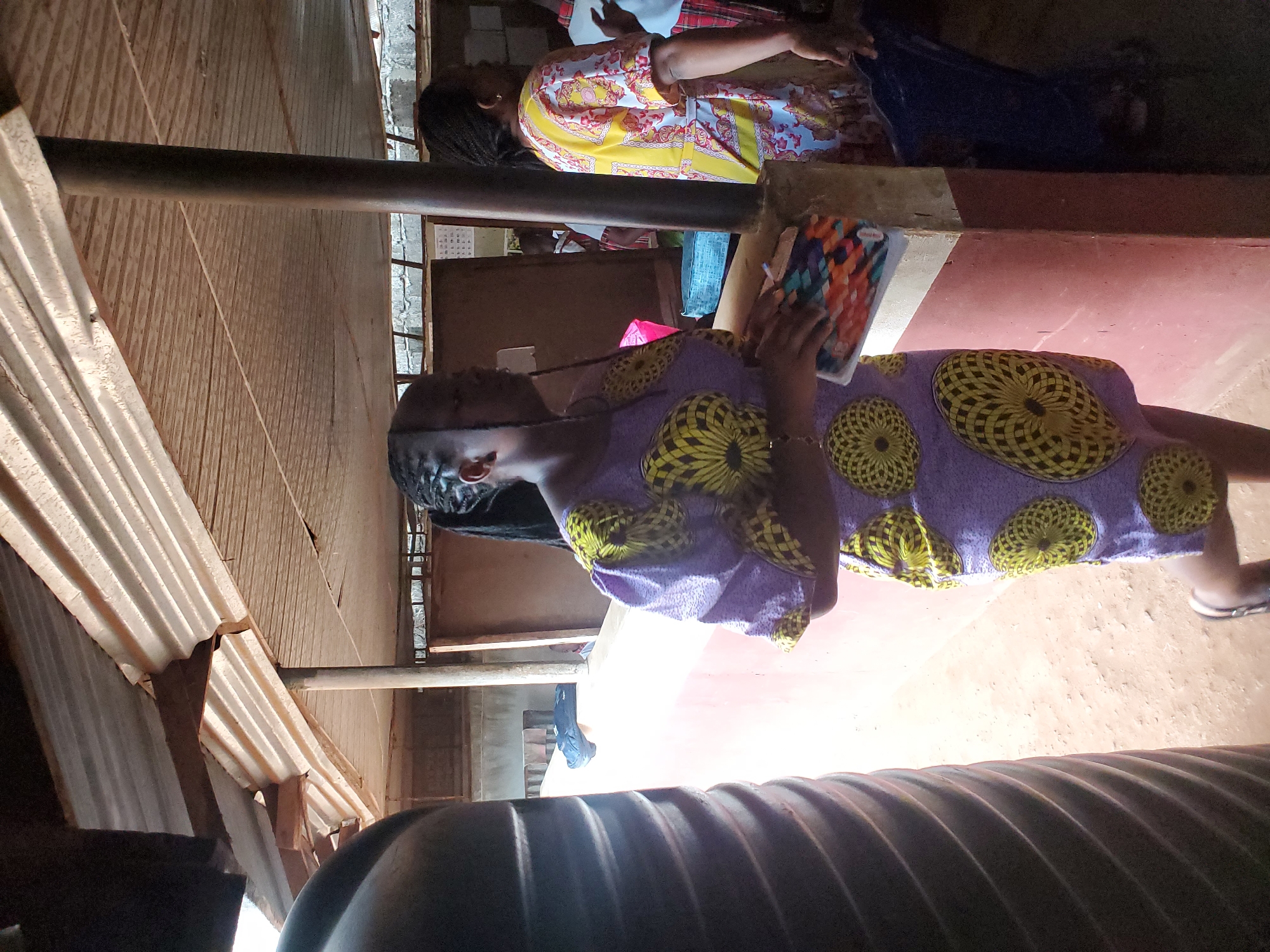 The image shows a scene inside what appears to be a modest structure in Ukwu-Abwa, Nigeria. Two women are visible. One is wearing a yellow and white patterned top with what appears to be blue trousers. The second woman, closer to the camera, is wearing a lilac dress with a yellow and black pattern, and she holds a brightly colored notebook. The ground is red, possibly made of compacted dirt or clay. The roof is corrugated metal. A steel pipe or beam runs horizontally across the middle of the frame.