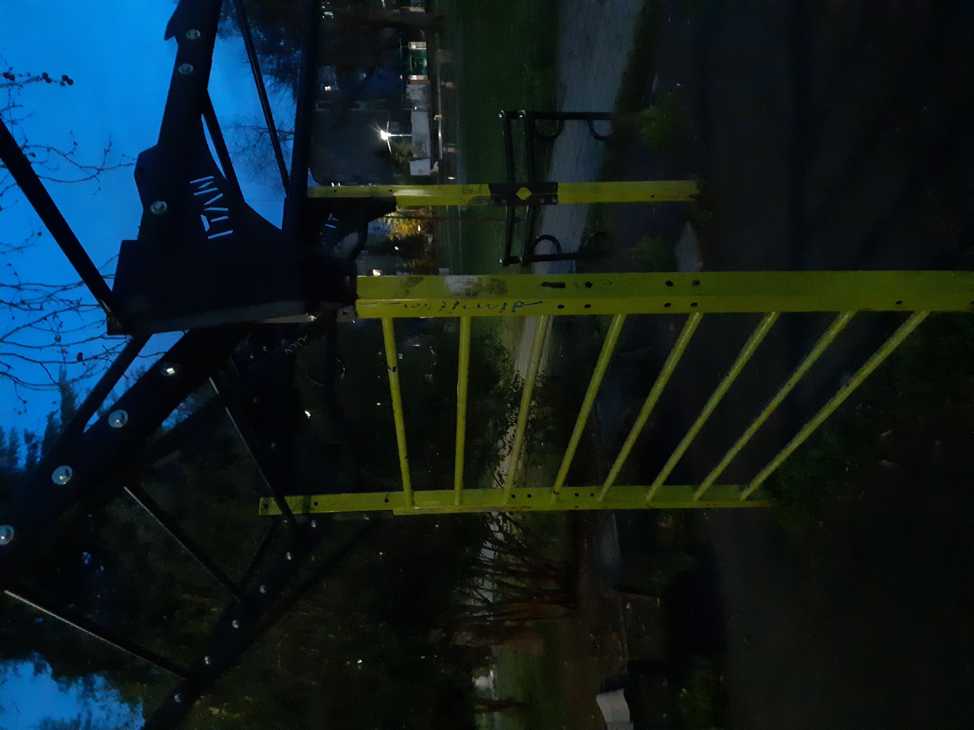 The image depicts a piece of exercise equipment located in Plovdiv, Bulgaria. The equipment features a black structure on the left with the word "IVAN" in white lettering. Adjacent to this is a yellow horizontal ladder. The background suggests a park setting with trees, a pathway, and distant buildings. The lighting indicates it is dusk or night. There is no visible human presence in the shot.