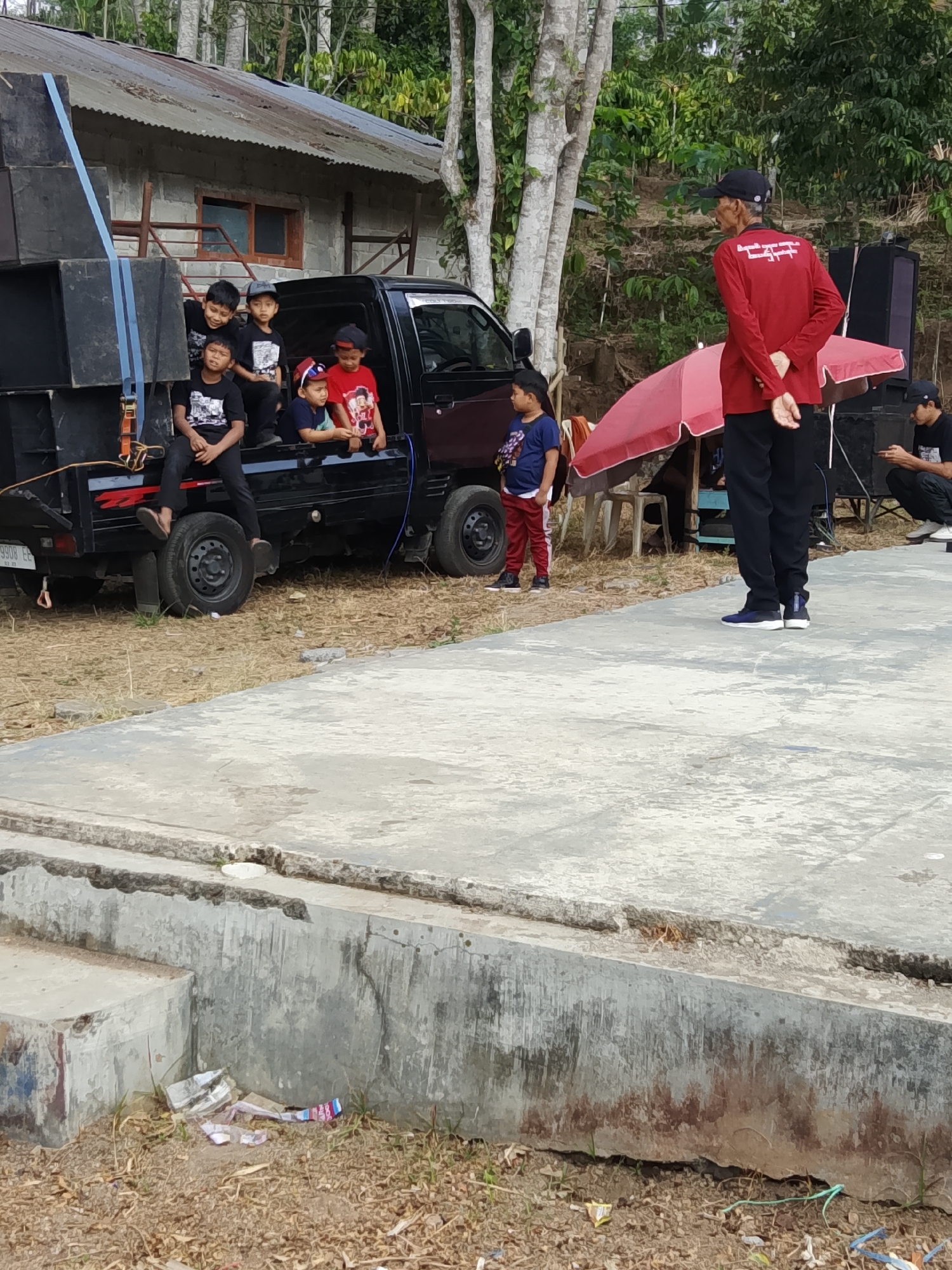In Jumo, Indonesia, a group of children are gathered around the bed of a black pickup truck, which is loaded with large speaker boxes. The truck is parked near a building with a corrugated metal roof and a stone facade. A man in a red long-sleeved shirt and black pants stands near a pink umbrella, facing away from the truck. Another man, wearing a black t-shirt and black pants, is seated in the background, looking down at his phone. The ground is covered with dry grass and dirt, and a concrete platform is visible in the foreground. The scene appears to be an outdoor gathering or event.