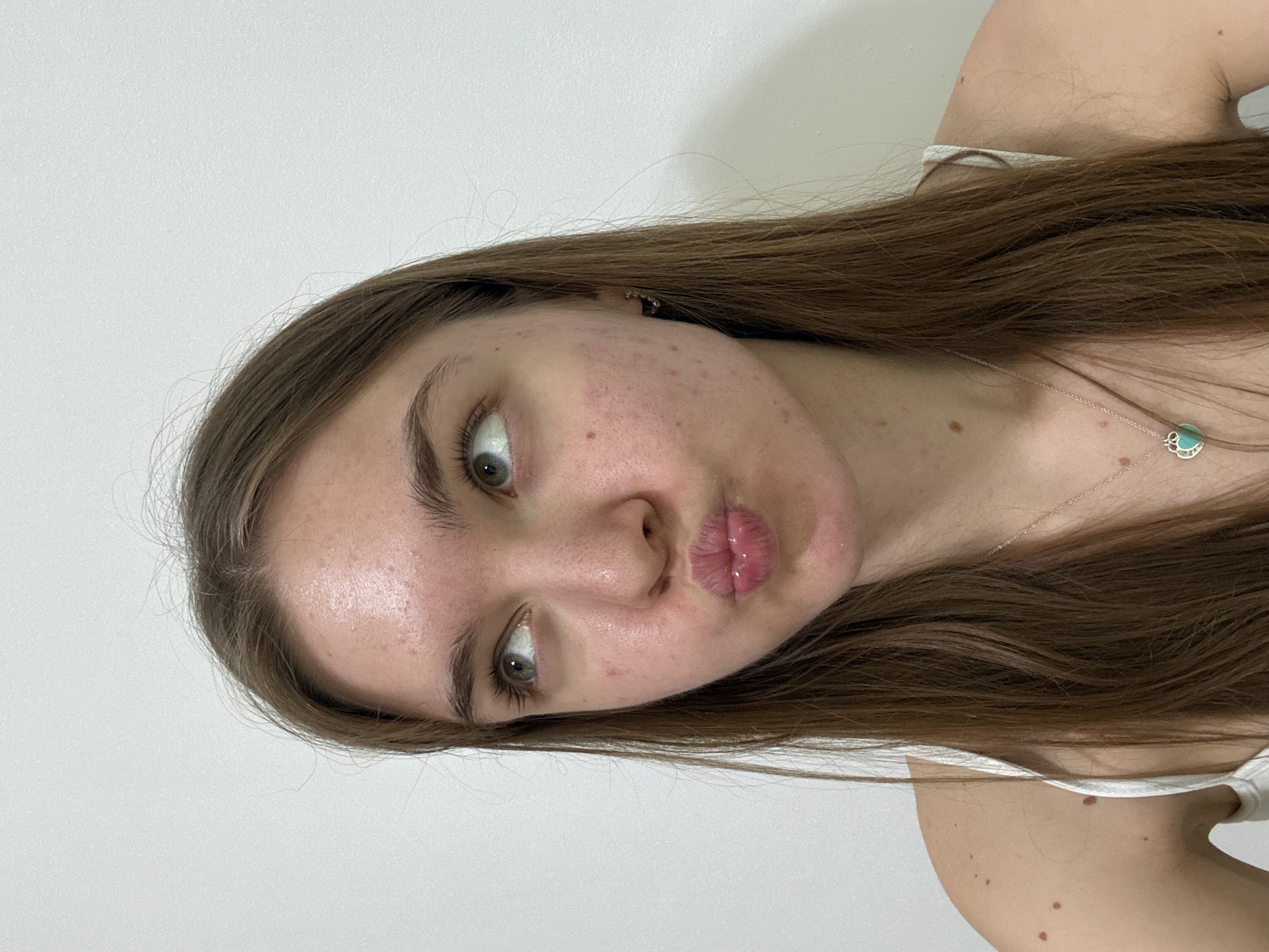 A fair-skinned female with brown hair is shown in a close-up, angled portrait against a plain white wall. She has green eyes and is making a kissing face. Her skin shows some acne and freckles. She is wearing a white top and a silver necklace with a blue pendant. No information about the city of Perm, Russia is relevant or visible in the image.