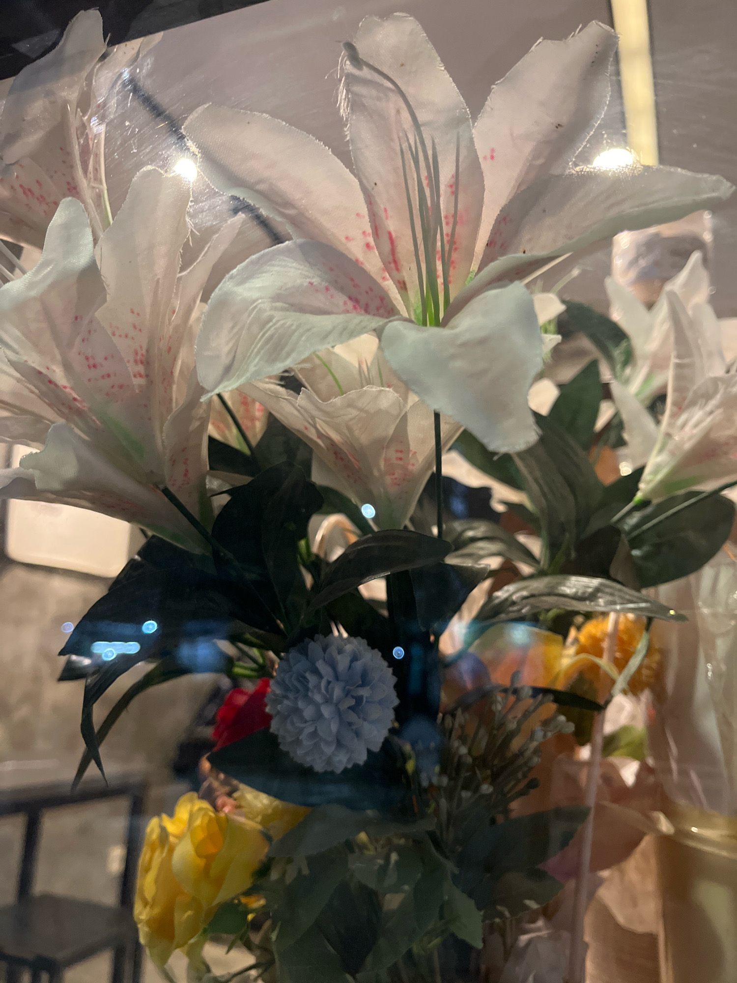 A close-up, low-angle shot shows a bouquet of artificial flowers. The prominent flowers are large, white lilies with pink speckles and thin green pistils. Behind them, dark green leaves create a backdrop. In the foreground and lower portion of the image, there are smaller flowers in shades of yellow, red, and a light blue pom-pom-like bloom. The bouquet is enclosed in clear plastic wrapping, which reflects light sources and the surrounding environment. The background is blurred and appears to be an indoor setting with muted lighting, possibly a commercial establishment, with glimpses of furniture and a textured wall. The overall impression is one of an artificial floral arrangement presented for display or sale. This image does not depict any identifiable location within Plajau Mulia, Indonesia, nor does it show any people, actions, or interactions.