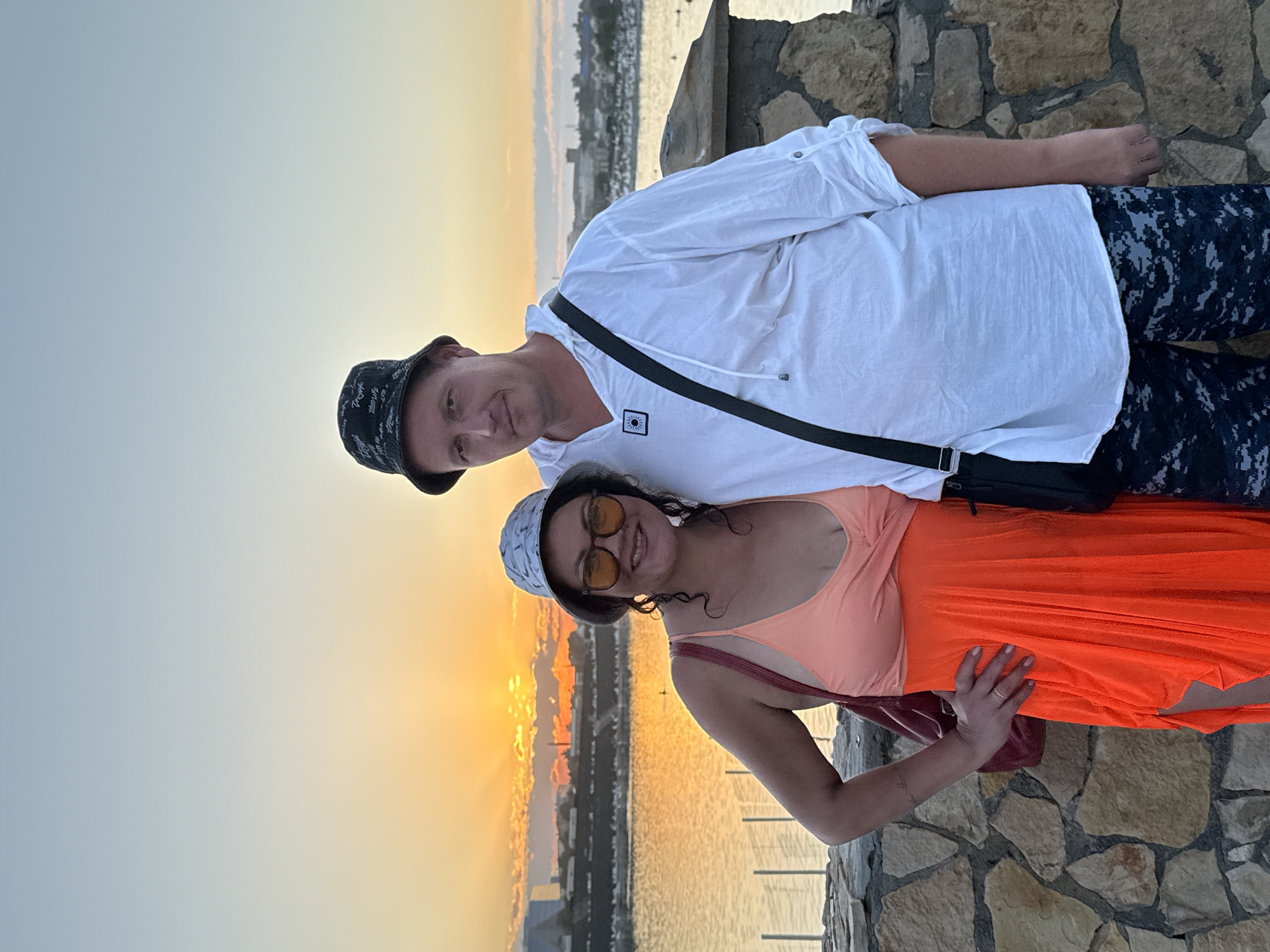 A man and a woman stand together outdoors in front of a body of water with a sunset in the background, which is likely the city of Sol-Iletsk, Russia, due to the distinctive stone embankment. The woman is on the left, wearing a bright orange dress and a light-colored bucket hat. She has dark, curly hair, glasses, and is smiling. She has her left hand on her hip and a maroon bag is slung over her right shoulder. The man is on the right, wearing a white long-sleeved shirt, a black bucket hat, and camouflage-patterned shorts. He has a black bag with a strap crossing his chest. They are both looking at the camera. The setting sun casts a warm orange glow across the sky and the water. The foreground features a stone wall.