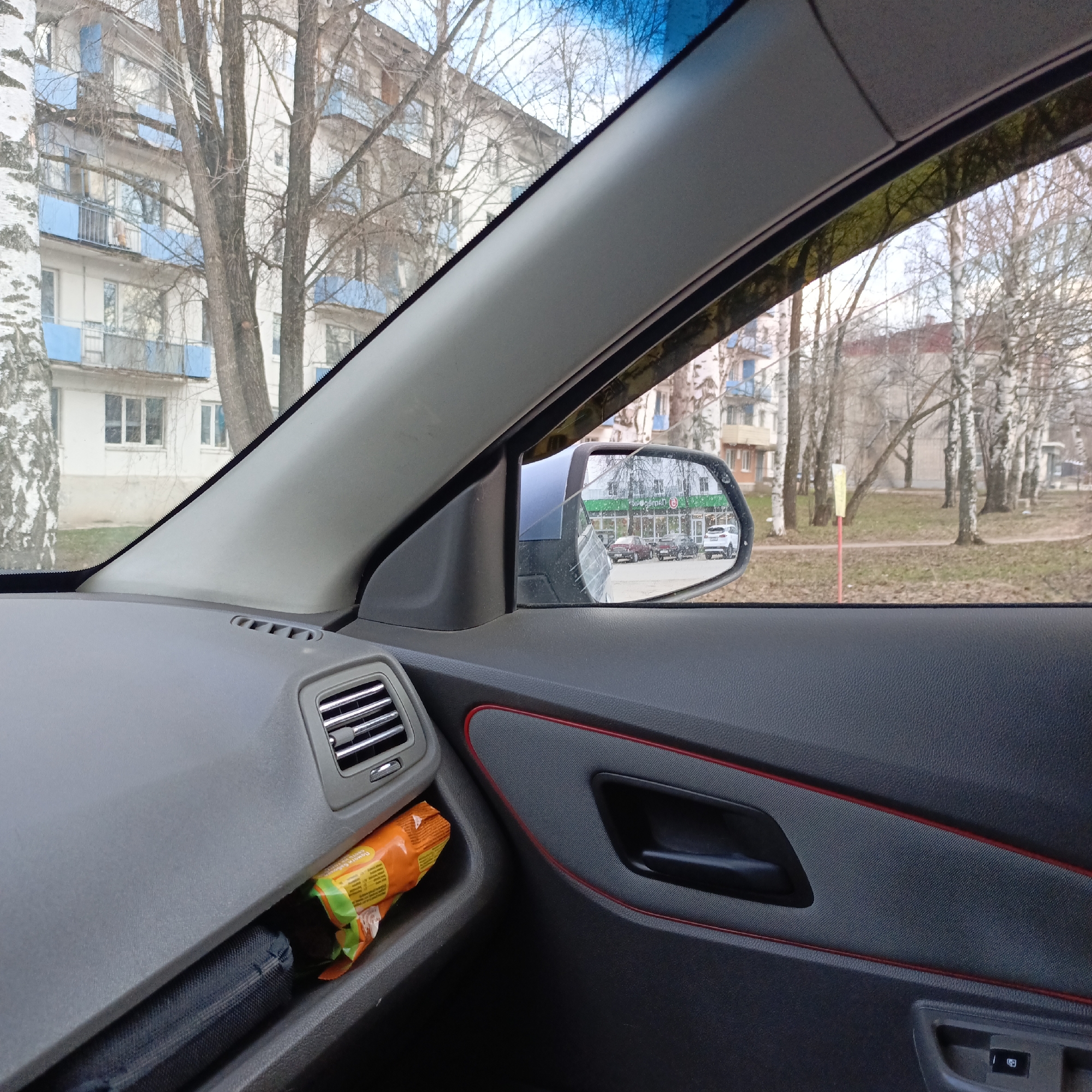 From the interior of a vehicle, the scene is of an urban area in Unknown, Unknown, observed through the window and side mirror. Outside the vehicle, there's a residential building with blue balcony railings, and trees, some of which are birch trees due to their white bark. The side mirror shows reflections of cars parked near a store with a green facade. Inside the vehicle, on the dashboard and door panel, are air vents, an orange snack package, a black case, and a door handle outlined in red. The overall ambiance suggests a cold, early spring or late autumn day.