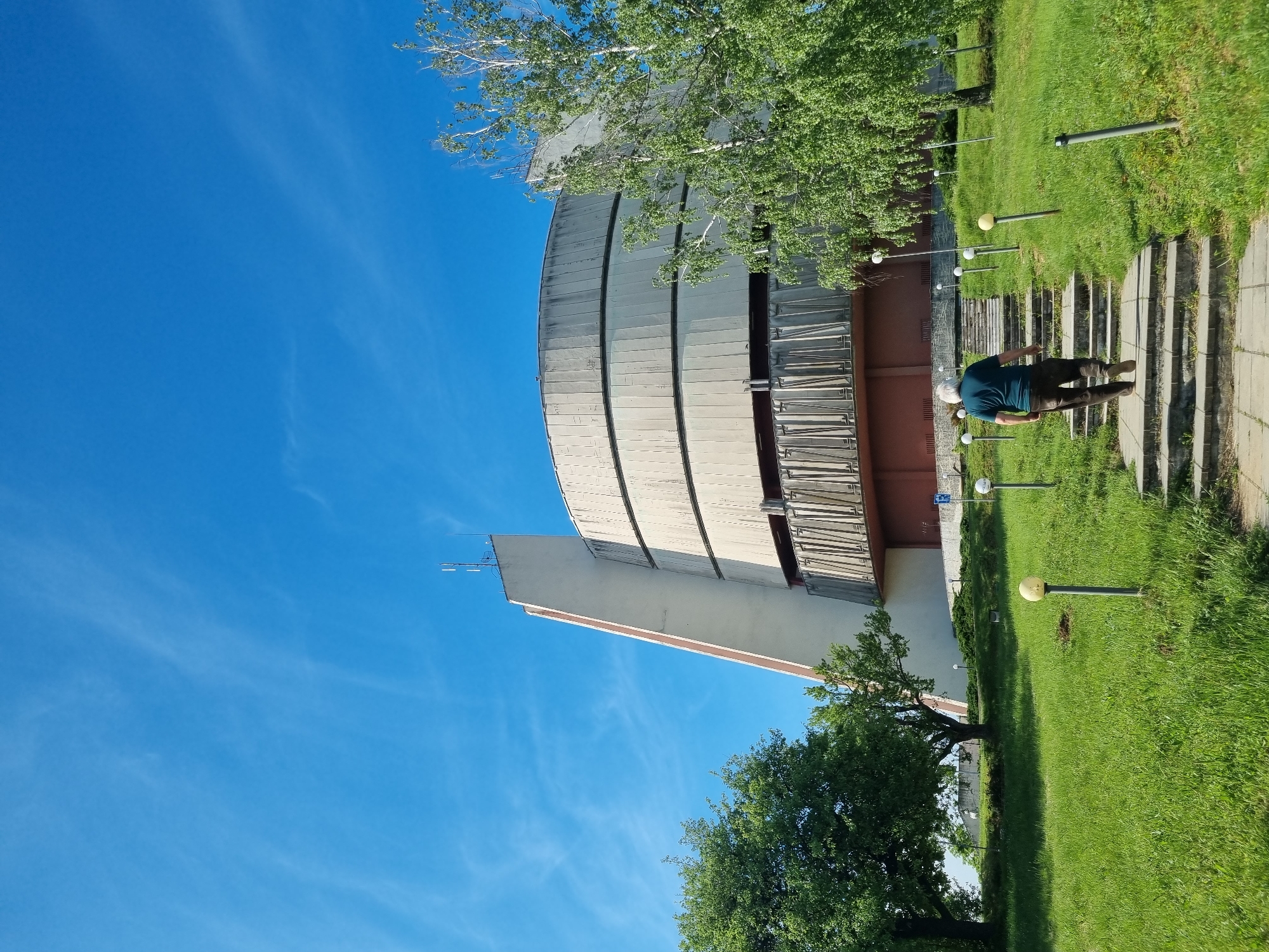 The image shows a tall, curved building with a cross atop a flat section. The building has a ribbed exterior. A man in a dark shirt and lighter pants walks up a series of concrete steps toward the building. The building is surrounded by a grassy area dotted with lamp posts with spherical lights. Trees are present. The sky is blue with wispy clouds. This appears to be the Pleven Panorama in Pleven, Bulgaria.