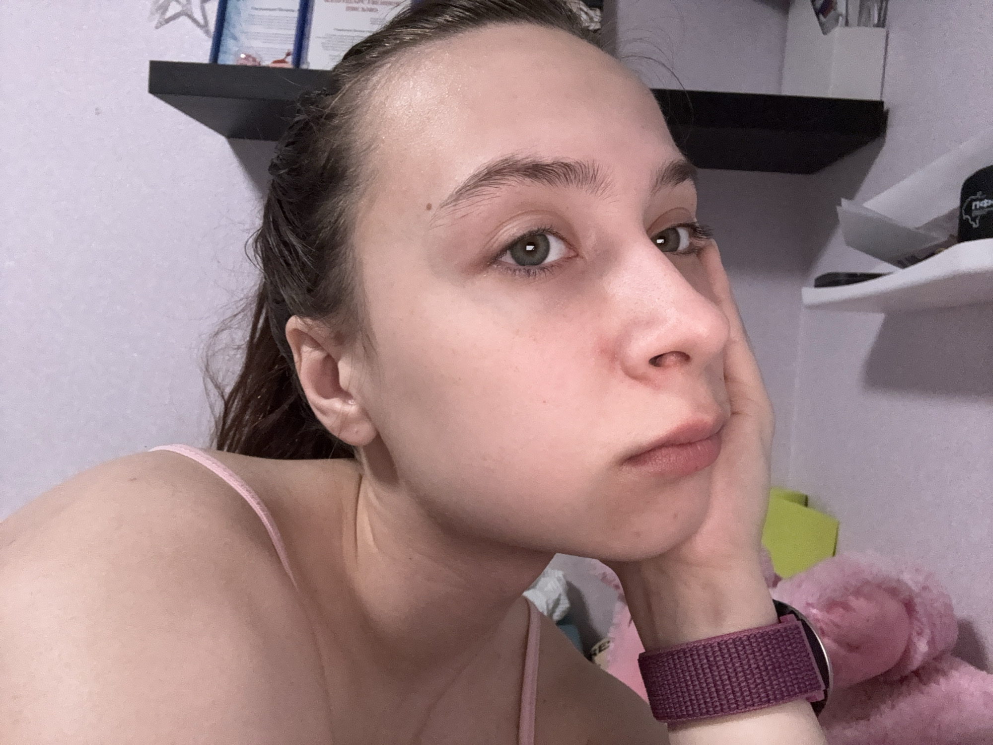 A young woman is positioned in the foreground, resting her cheek on her hand. She has fair skin, brown hair pulled back from her face, and light green eyes. She is wearing a pink tank top and a purple watch.
Behind her, there are two shelves mounted on a light purple wall. Items are visible on the shelves, including a white container. A pink stuffed animal is also in the background.
The location of Прикамский, Russia is not visibly indicated in the image.