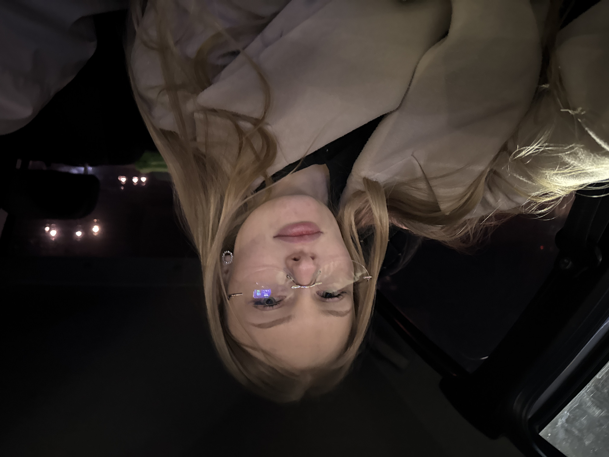 A young woman with blonde hair is in the driver's seat of a car, possibly looking up at the car's sunroof. She is wearing frameless glasses and earrings. The car is dark inside, but there are blurry lights visible through the windshield. Given the prompt, the location is possibly Perm, Russia.
