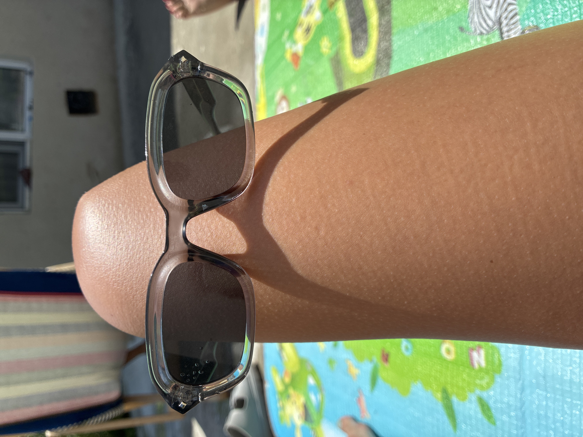 The image shows a close-up view of a person's leg, which is resting on a colorful mat. The leg is tanned and appears to have goosebumps. A pair of sunglasses with transparent frames is placed on the leg. In the background, a striped beach chair and a building with windows are visible. The scene appears to be outdoors on a sunny day. The location is not specified in the image itself or the provided crops.