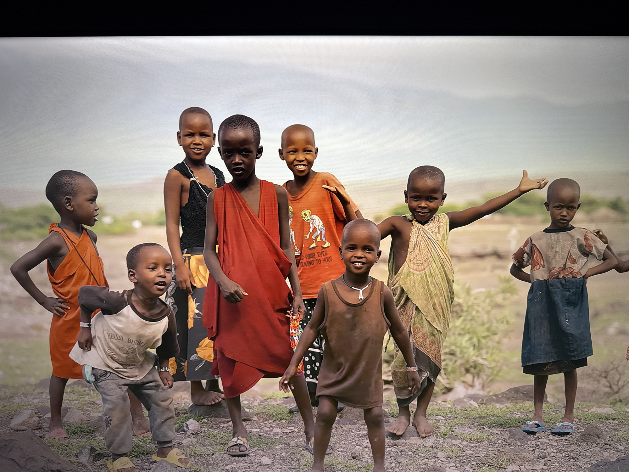 A group of nine children, varying in age, stand together outdoors. The background suggests a rural or natural environment with a cloudy sky and some distant foliage and structures. The children are positioned in a staggered arrangement, facing towards the viewer, with some making eye contact. Their clothing varies, with some wearing traditional garments and others in more casual attire. One child in the foreground is wearing a brown tank top and pants, with a necklace. Another child to their left wears an orange dress. A child in the center wears a red wrap. To the far left, a child is dressed in an orange tunic. Further back, another child wears a black top and a beaded necklace. A child in an orange t-shirt with a graphic print is visible in the middle. To the right, a child in a patterned wrap stands next to a child in a blue dress. The ground beneath them appears to be dirt and rocks. The lighting is diffused, indicating an overcast day.