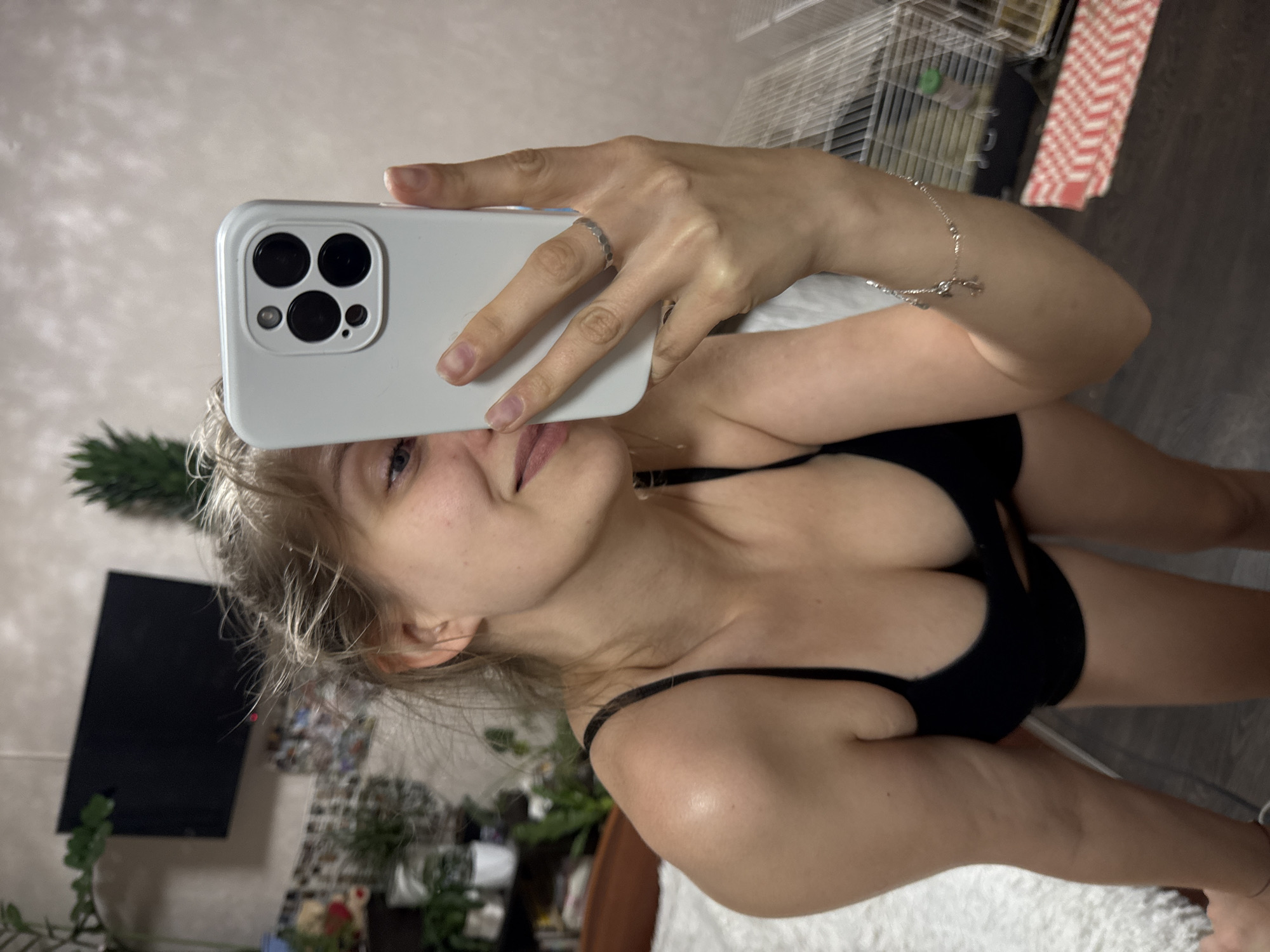 A young woman with blonde hair tied back in a messy bun is taking a selfie in a bedroom. She is wearing a black bra and shorts. She is holding a white smartphone in her right hand. The background includes a bed with white bedding, a shelf with plants, a television on the wall, and a birdcage. The room appears to be dimly lit.