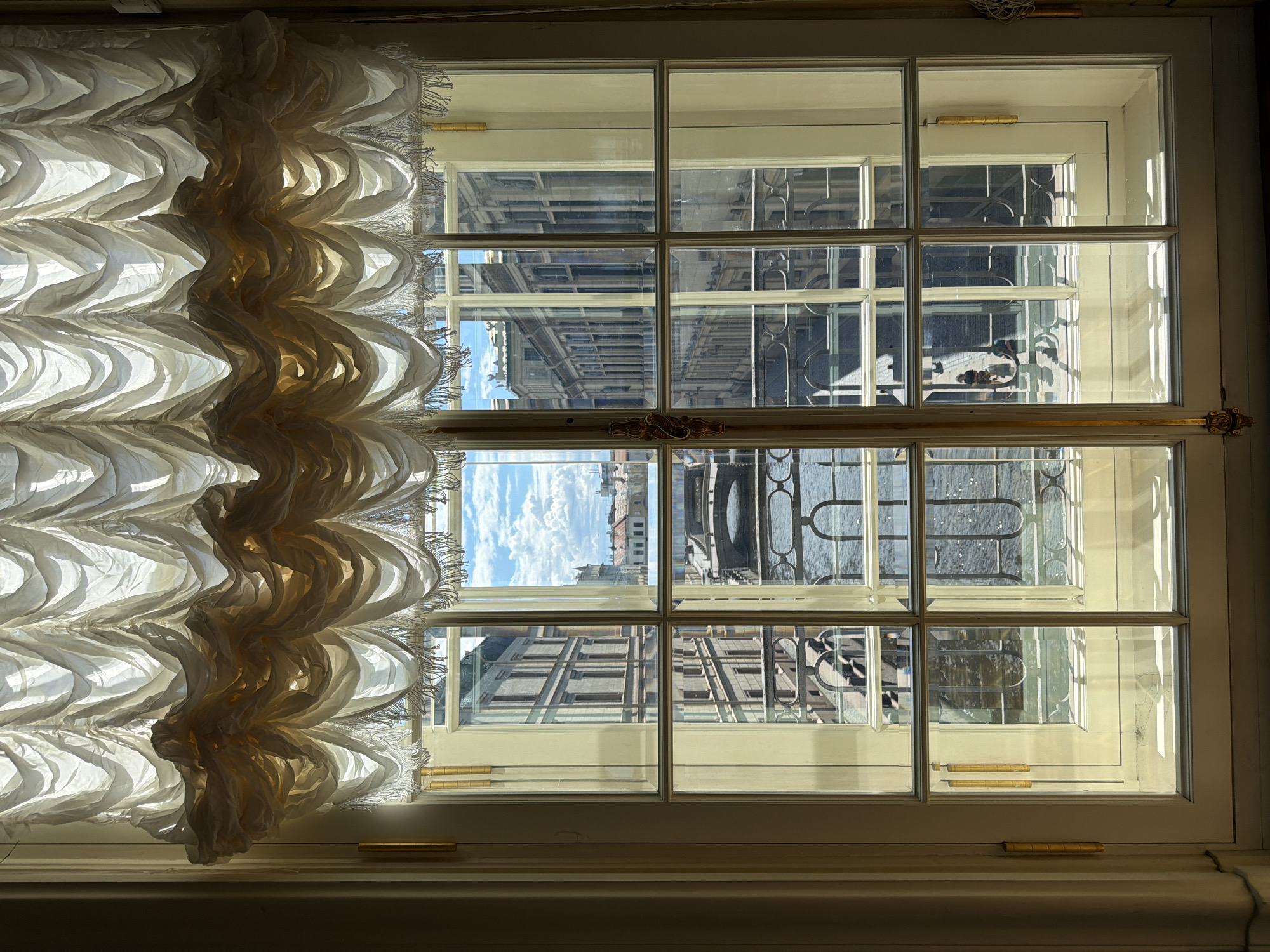 A close-up shot frames a large window with multiple panes. A decorative curtain, predominantly white with some beige accents, drapes across the left side of the window, its fabric gathered and ruffled, creating a textured and voluminous appearance. The curtain has a fringe along its top edge, with individual threads visible.
Through the window, a city view is partially visible. The panes reflect the sunlight, creating bright areas and obscuring some details. In some panes, the architecture of buildings is discernable, with rows of windows and patterned railings. The sky, with scattered clouds, is also visible through certain sections of the window, suggesting it is daytime. The architectural style and overall impression evoke a European city, consistent with the Saint Petersburg, Russia location context. The light suggests a bright day, possibly morning or afternoon. No text is visible in the image. The scene conveys a sense of quiet observation, looking out from indoors onto an urban landscape.