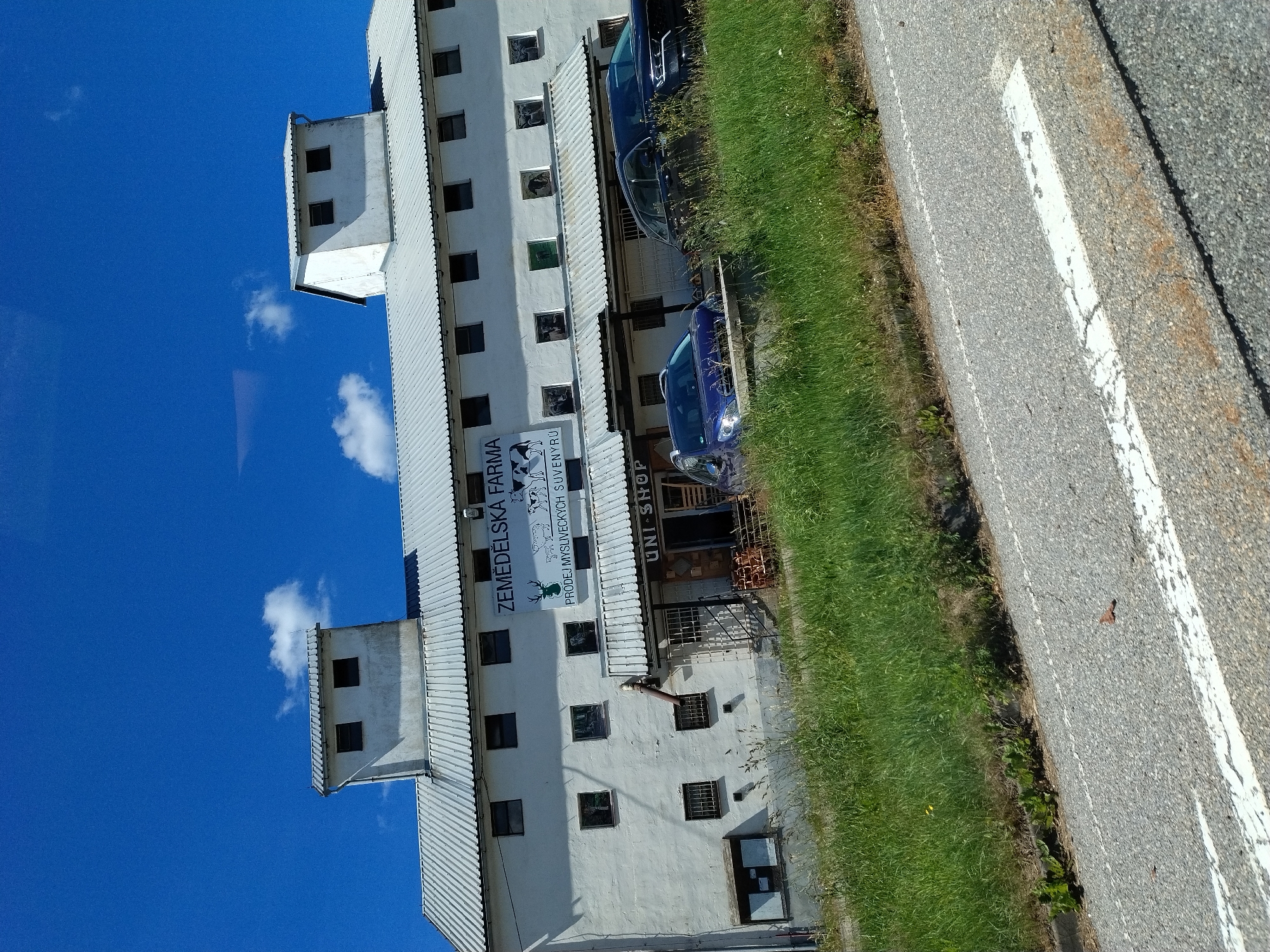 The image shows a white, multi-story building with a corrugated metal roof, identified by a sign as "ZEMEDELSKA FARMA" (Agricultural Farm). The sign also indicates "PRODEJ MYSLIVECKYCH SUVENYRŮ" (Sale of Hunting Souvenirs). The building has numerous windows and two distinct tower-like structures on the roof. In front of the building, there is a covered area with a sign that reads "UNI-SHOP." Two cars are parked in front of the shop, a blue hatchback and a dark SUV. The building is situated alongside a grassy embankment and a paved road. The sky is bright blue with a few scattered clouds. The image appears to have been taken from a vehicle moving along the road.