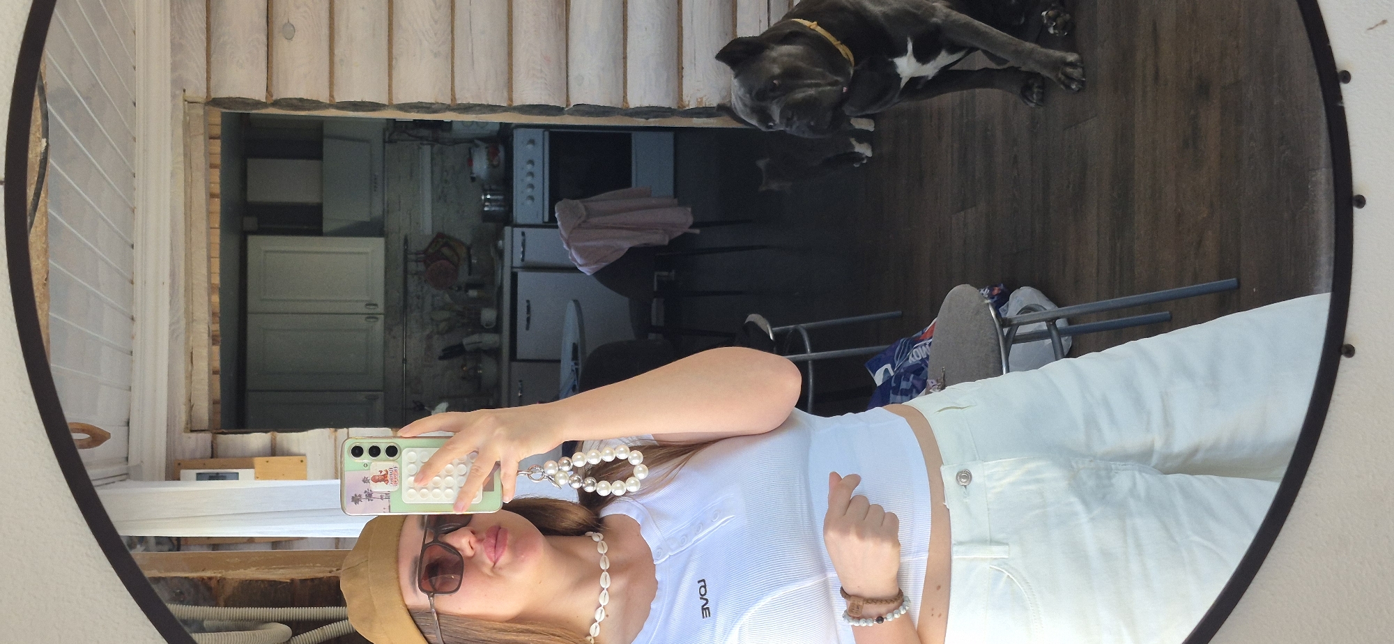 The image depicts a selfie taken in a mirror, likely in a residential setting. A woman, wearing a white top, white pants, a tan hat, and sunglasses, is holding a smartphone to capture the image. Her phone has a light green case and a beaded phone charm. She is also wearing a shell necklace and bracelets. The mirror reflects a kitchen and dining area. In the background, there is a large, dark-colored dog with white markings lying on the floor. The location is within a house with wooden walls. The specific city is not discernable from the image.