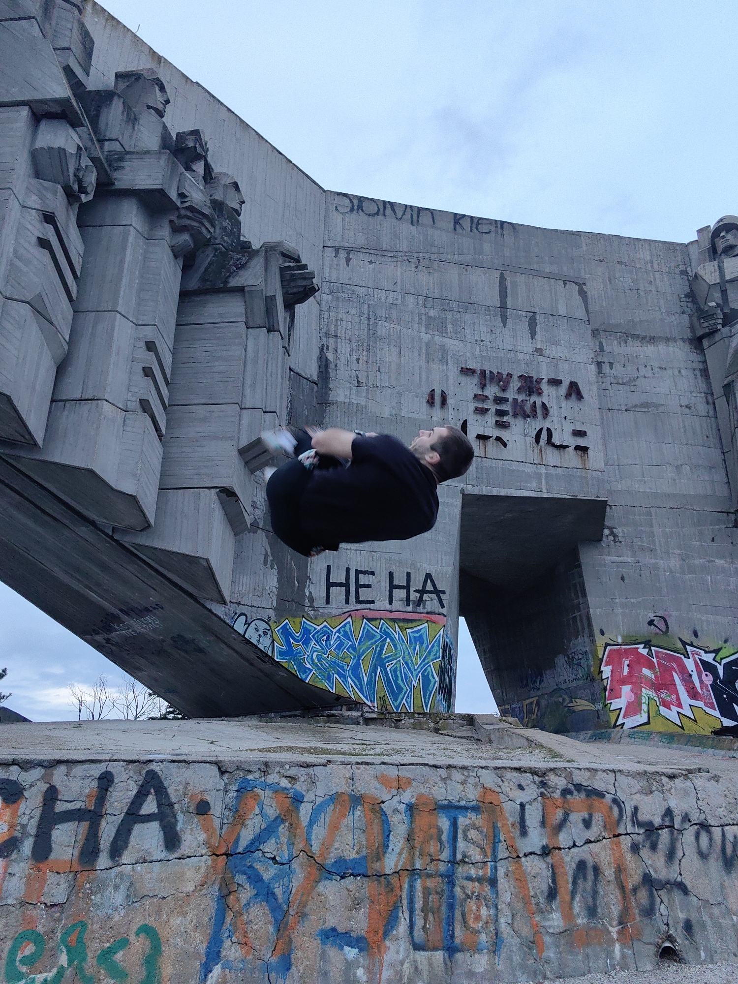 A man is performing a backflip in front of the Monument of the Bulgarian-Soviet Friendship in Varna, Bulgaria. The monument is a large, concrete structure with carvings of faces and Cyrillic script. Graffiti covers the monument's base. The sky is overcast.
