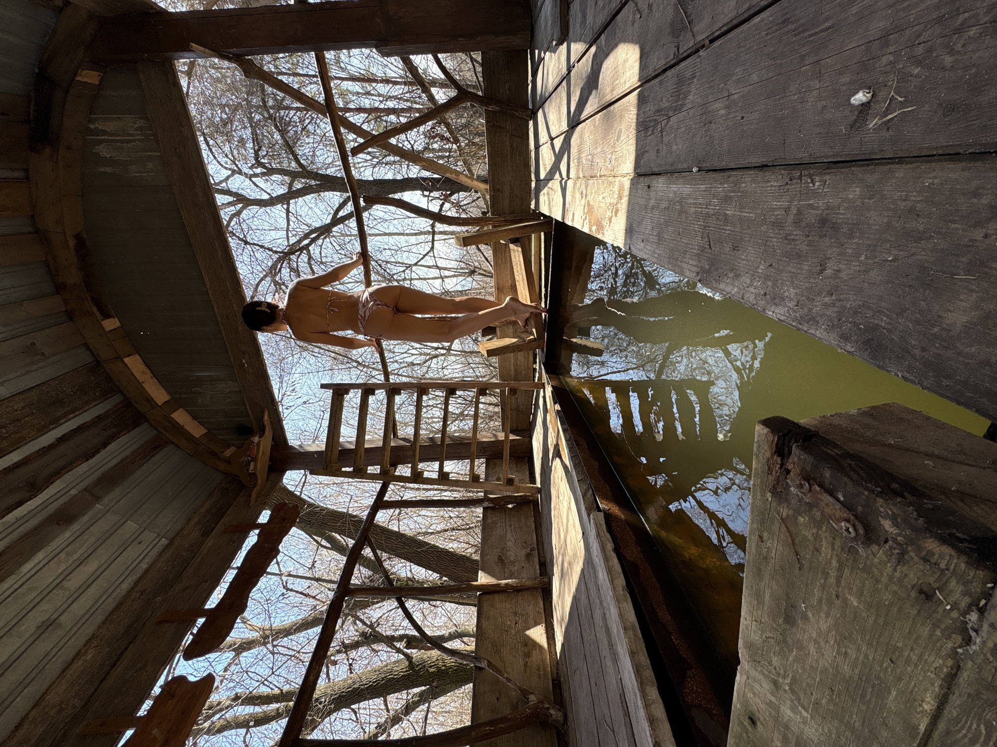 A low-angle shot depicts a woman standing on a wooden platform above a body of water, likely a lake or pond, within what appears to be a rustic wooden structure. The woman is wearing a bikini and holding onto a wooden pole. A wooden ladder leads up to the platform. The structure seems to be partially enclosed with corrugated metal sheeting on one side. Bare tree branches are visible through gaps in the structure, suggesting it is located in a rural or semi-rural environment. The water surface reflects the sky and surrounding structures. The scene has a rough, aged appearance, with weathered wood and visible construction details. The location is within Ulyanovsk, Russia.