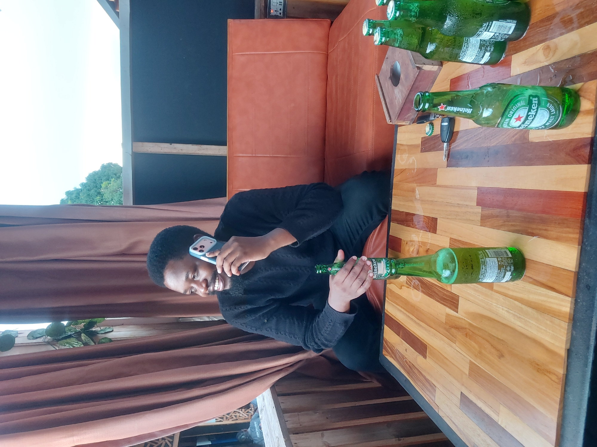 A man is sitting at a table with multiple bottles of Heineken beer. He is holding one bottle and talking on a mobile phone. A car key is on the table. The location appears to be an outdoor seating area. The city is not possible to be determined from this image.