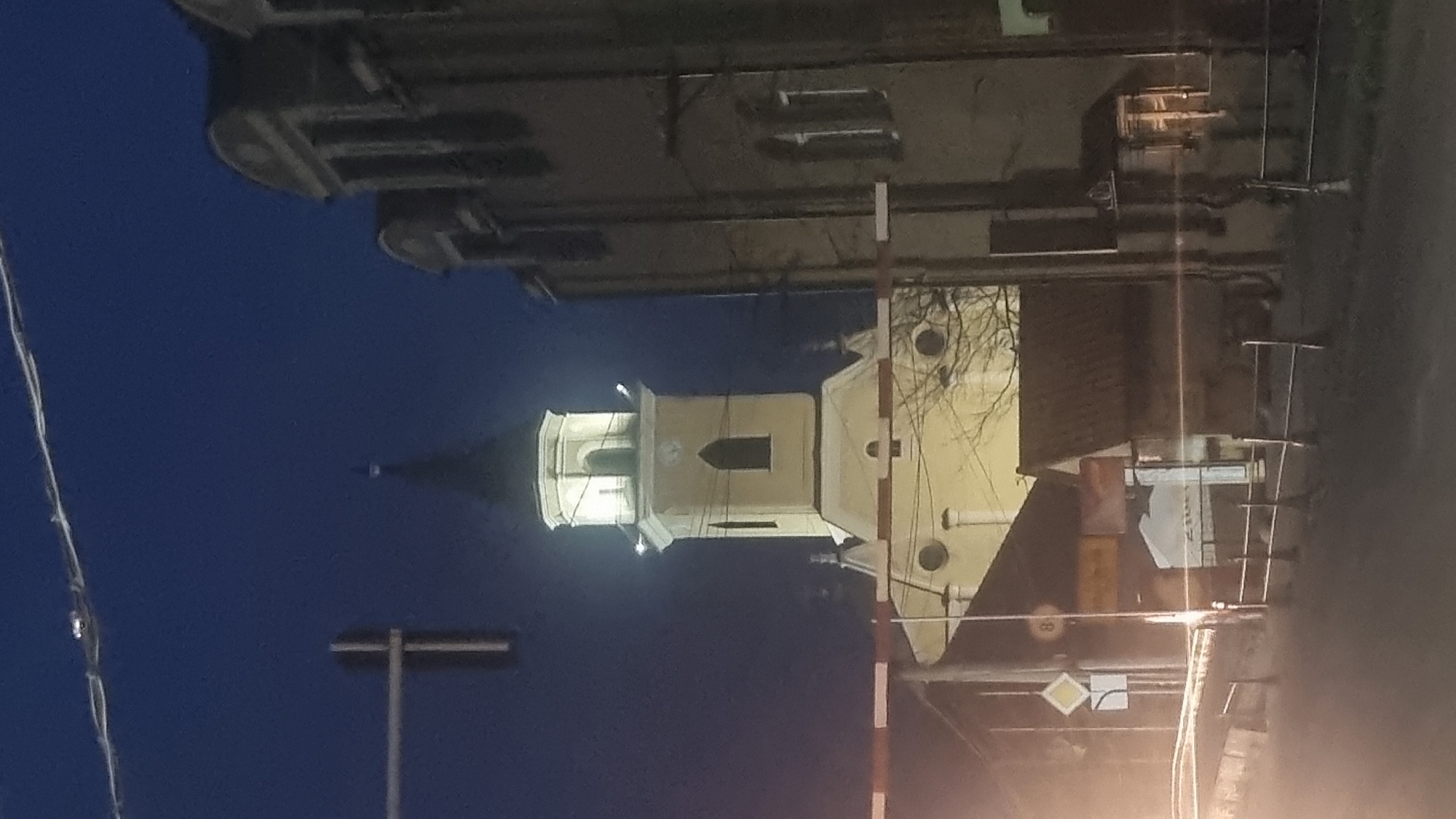 The image depicts a night scene in Berehove, Ukraine, focusing on a church tower and adjacent buildings. The sky is a deep, dark blue. The prominent church tower is light yellow with a black spire and is illuminated from within. It has a clock face and arched windows. To the right of the church, a darker, multi-story building with arched windows is visible. In the foreground, there are street elements including a railway crossing barrier, a traffic light pole, and fences. The ground appears to be a road. The overall impression is an urban night view with a notable architectural landmark.