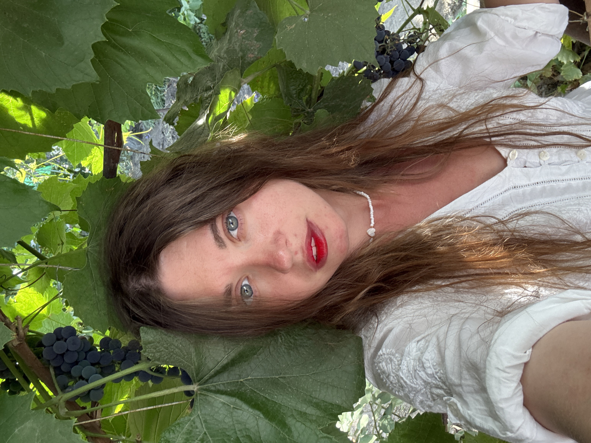 A woman with light skin, blue eyes, and long brown hair is taking a selfie outdoors. She is wearing a white embroidered shirt and a white beaded necklace. She has bright red lipstick on. Behind her are large green grape leaves and dark purple grapes on a vine. The scene appears to be in a garden or vineyard, potentially in the city of Sol-Iletsk, Russia. The lighting suggests it is daytime, with sunlight filtering through the leaves.