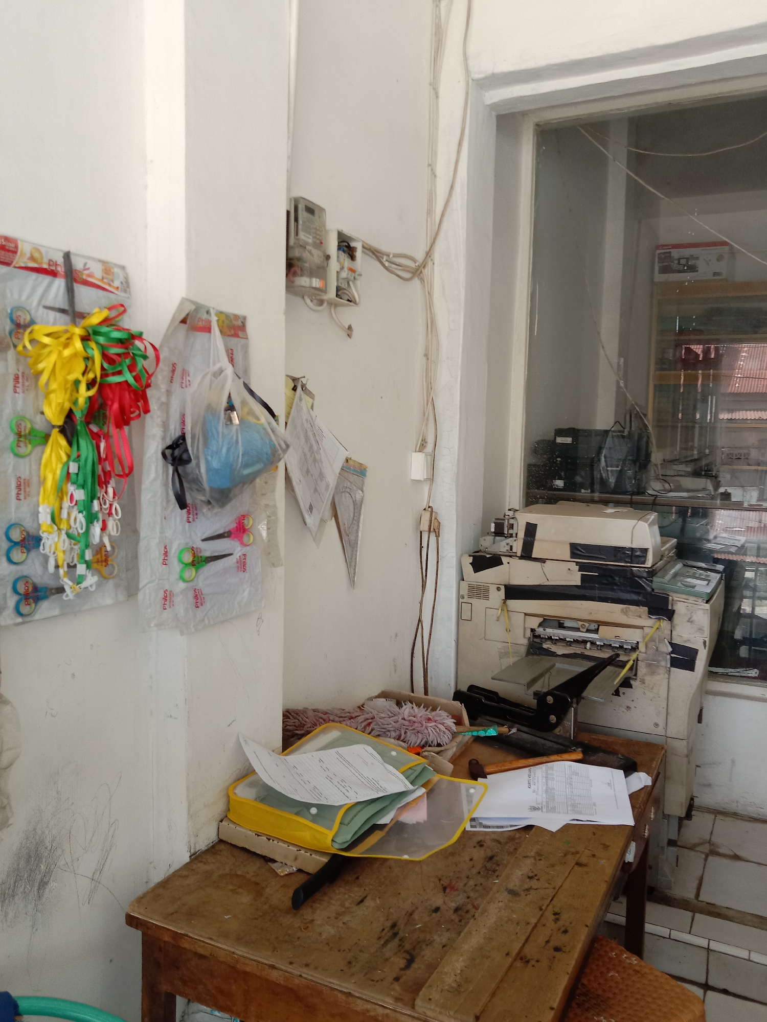The image depicts an indoor scene in a room, likely in Lhokseumawe, Indonesia. The room features a worn wooden desk cluttered with papers, a yellow document folder, a hammer, and cleaning supplies. An old, taped-up photocopier sits near a window. On the wall, there are packages of scissors and ribbons hanging, along with electrical meters and wiring. The environment appears disorganized and utilitarian. There are no people visible in the image.