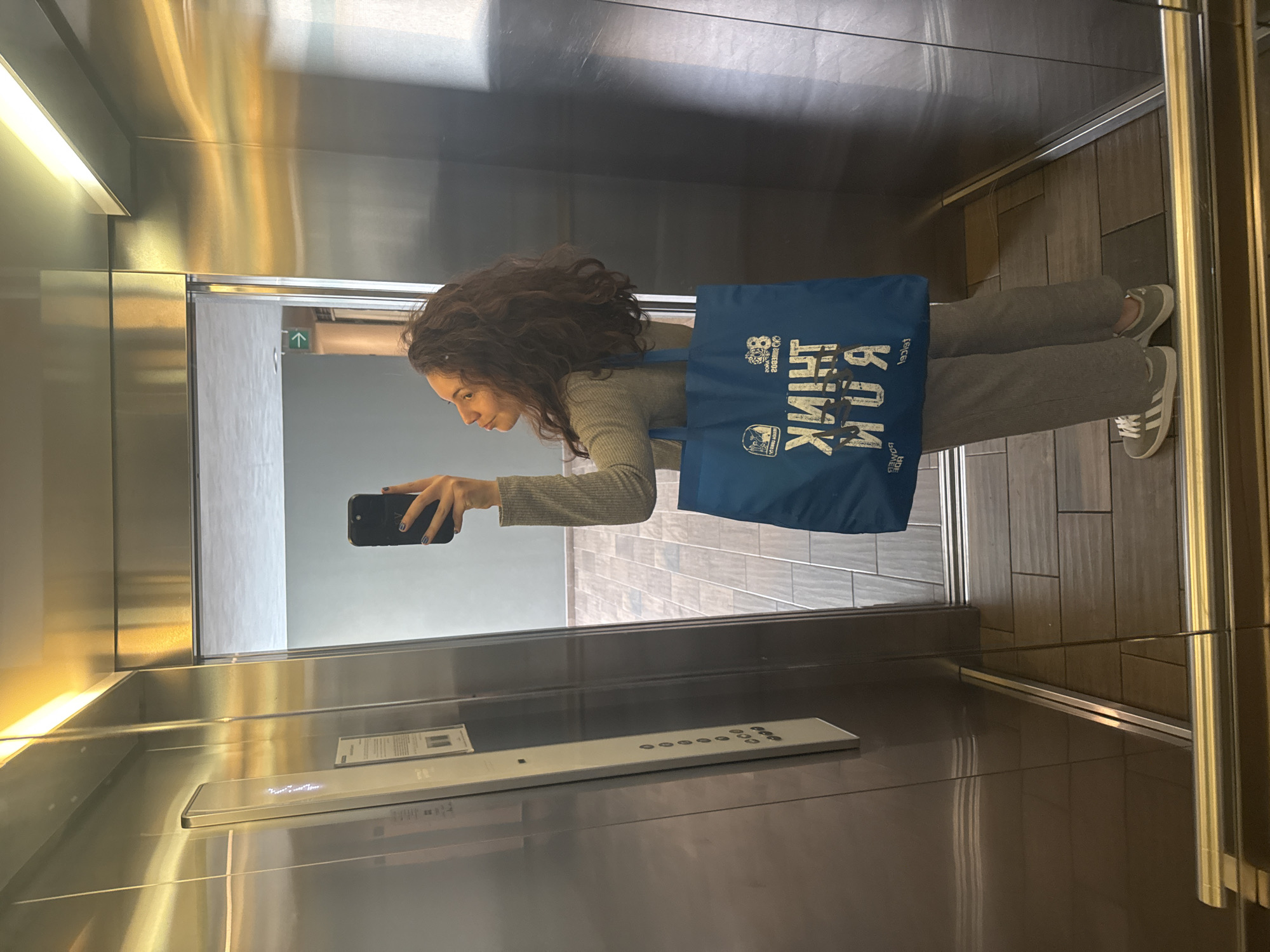A woman with dark curly hair takes a selfie in an elevator. She wears a light-colored, long-sleeved shirt and gray pants with gray Adidas sneakers. She is holding a black smartphone in her left hand. She is also wearing a blue tote bag with white lettering. The elevator interior has metallic walls and a tiled floor. In the background is the reflection of a wall with light gray tiles. The city is not visible in this image.