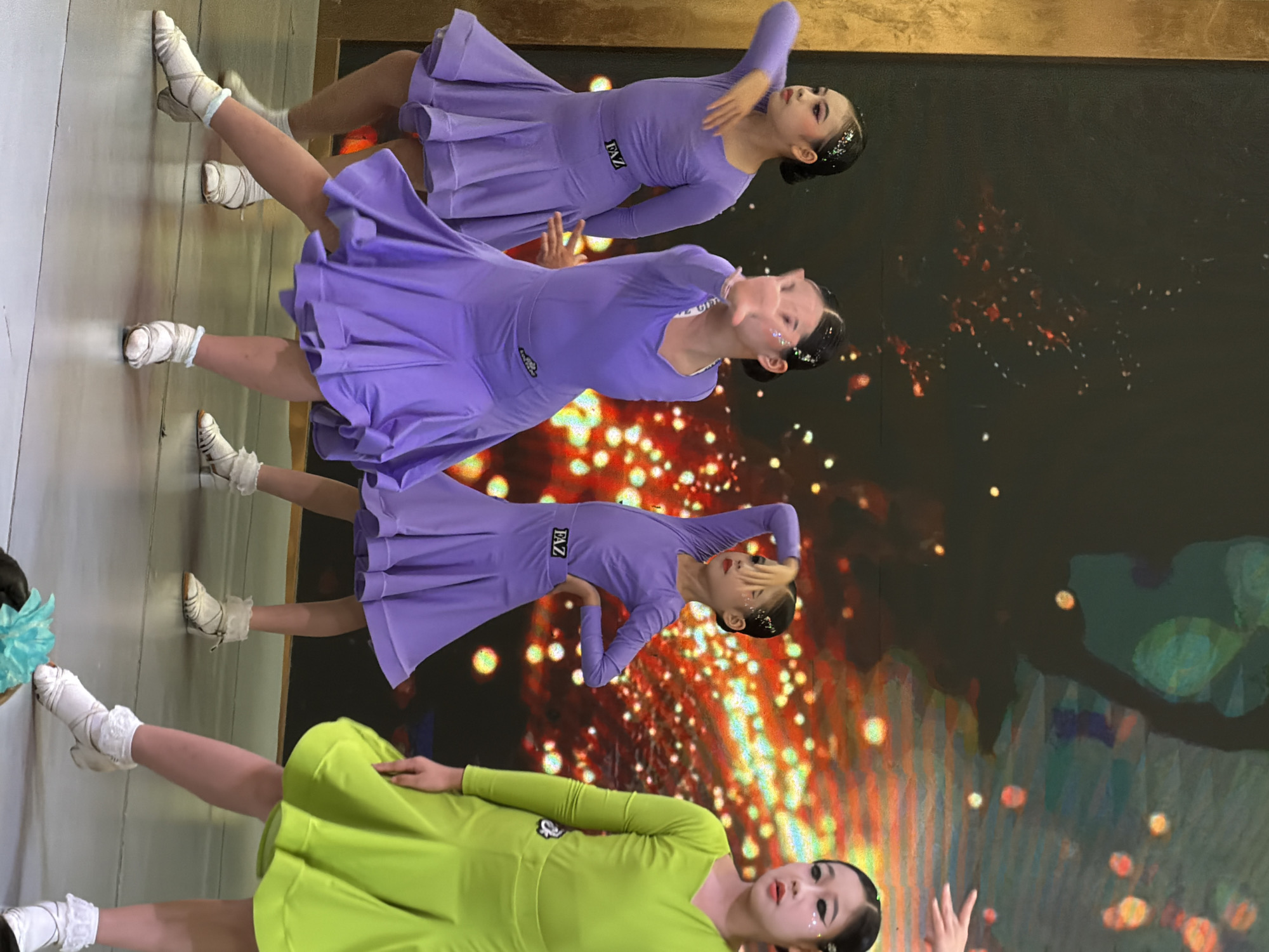 In Jiulongpo District, China, four young girls in matching purple dance outfits perform on a stage. The girl on the far left is positioned facing the left, with her left arm extended and bent at the elbow, while her right arm is bent and held in front of her body. She wears a purple dress with a flared skirt and white socks. To her right, another girl in a similar purple dress is captured mid-pose, with her right arm extended towards the viewer and her left arm bent. Her gaze is directed forward. Further to the right, a third girl in a purple dress strikes a similar pose, with her left arm extended and bent, and her right arm in front of her. Her hair is styled with glitter. The girl on the far right wears a bright green dance dress. The stage floor appears to be a reflective metallic surface. Behind the performers, a large screen displays a vibrant, abstract pattern of colorful lights and shapes. The overall scene depicts a dance performance or rehearsal.