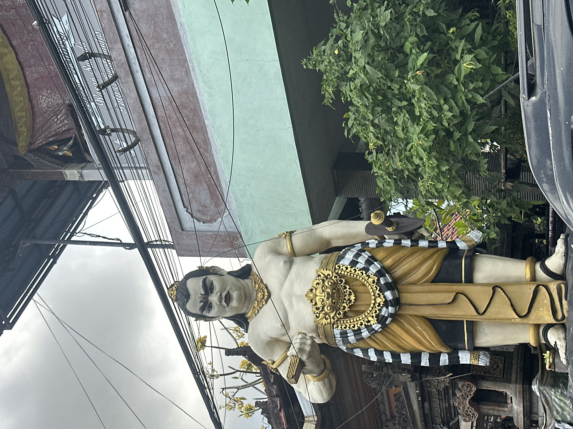 This is a medium shot, eye-level, outdoor, daylight photograph of a statue in Denpasar, Indonesia. The statue, which appears to be made of painted concrete, depicts a muscular, shirtless male figure with dark hair pulled back, wearing a golden crown, a thick golden necklace, a golden belt with ornate detailing, and a gold and black sarong. He is holding a rectangular object in his left hand and a sword-like object in his right hand. The statue is positioned in front of a building with a light green lower facade and a red-brown upper facade, with a dark corrugated roof structure overhead. Electrical wires are visible crisscrossing the scene. To the right of the statue, a large, lush green tree with yellow flowers is prominent. In the foreground, the roof of a dark-colored vehicle is visible. The sky is overcast and cloudy. The building appears to have a sign with some text, possibly in Indonesian, that reads "Wayan Suba" and "Fatu".