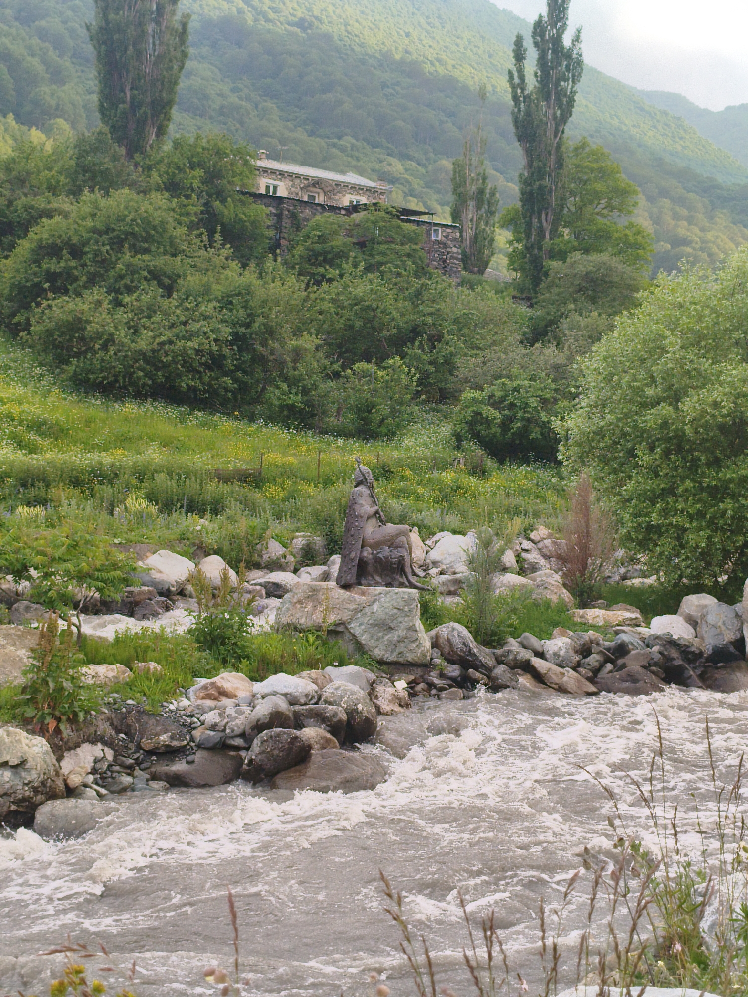 The image depicts a natural landscape with a focus on a fast-flowing, muddy river in the foreground. The riverbank is lined with large, grey rocks and interspersed with greenery. A bronze statue of a seated figure is positioned on a rock formation near the river's edge. Behind the river and statue, a lush green hillside rises, dotted with trees. A stone building with a tiled roof is visible on the hillside, partially obscured by foliage. Tall, slender trees stand prominently against the backdrop of a misty, forested mountain range. The overall scene suggests a tranquil, somewhat remote natural setting. While the prompt specifies Verkhny Fiagdon, Russia, there are no direct visual cues within the image to confirm this specific location.