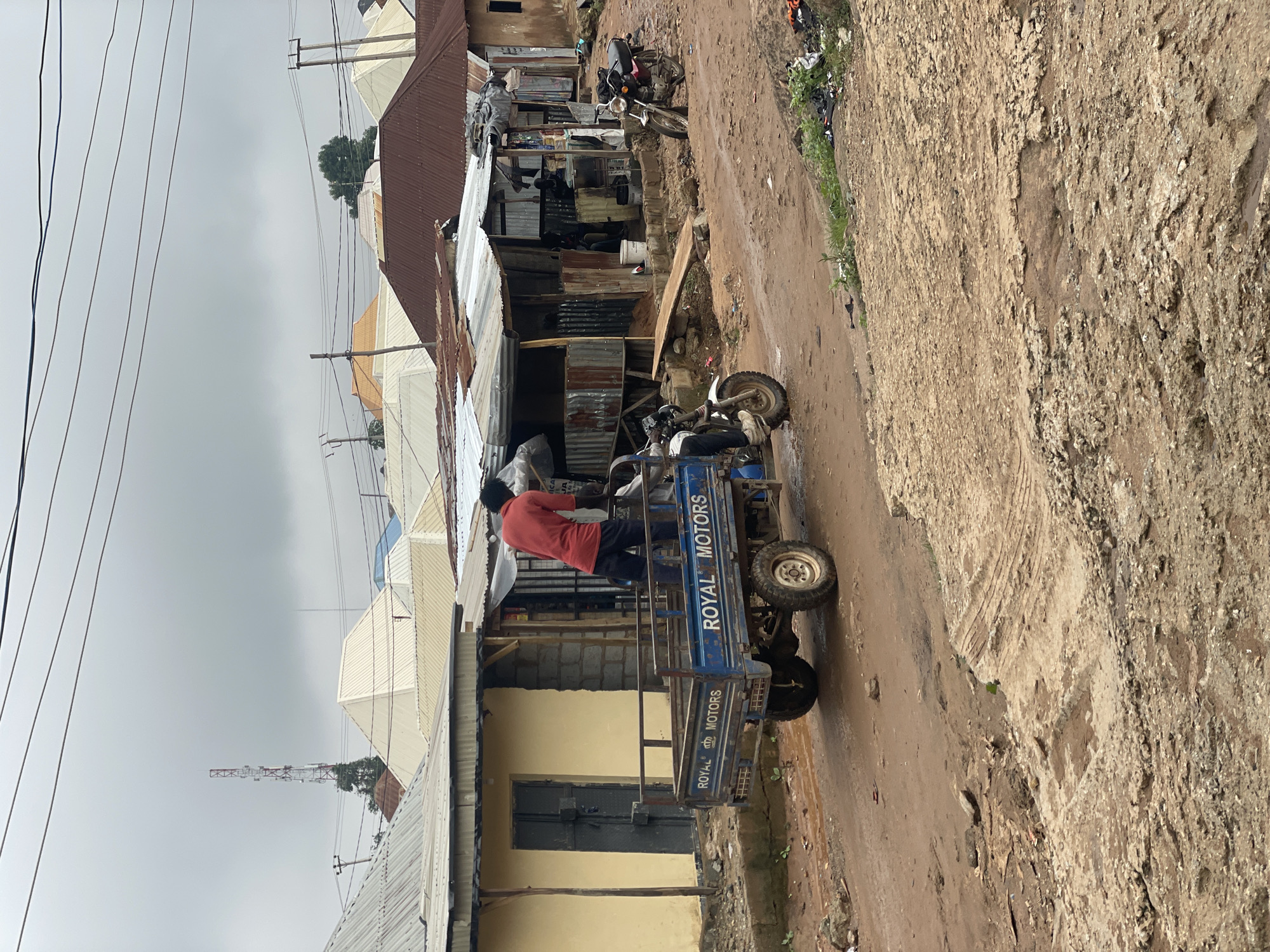 This image depicts a street scene in Kurunduma, Nigeria. A person wearing a red shirt and dark pants is standing in the back of a blue three-wheeled vehicle, appearing to be loading or unloading items. The vehicle has "ROYAL MOTORS" written on its side. The surrounding environment is characterized by unpaved, muddy roads with visible tire tracks and some debris. Several buildings with corrugated metal roofs and some with yellow painted walls line the street. In the background, a tall telecommunications tower is visible against an overcast sky, along with numerous residential or commercial structures with varied roof designs. A motorcycle is parked on the right side of the road, and vegetation is present in patches along the roadside.