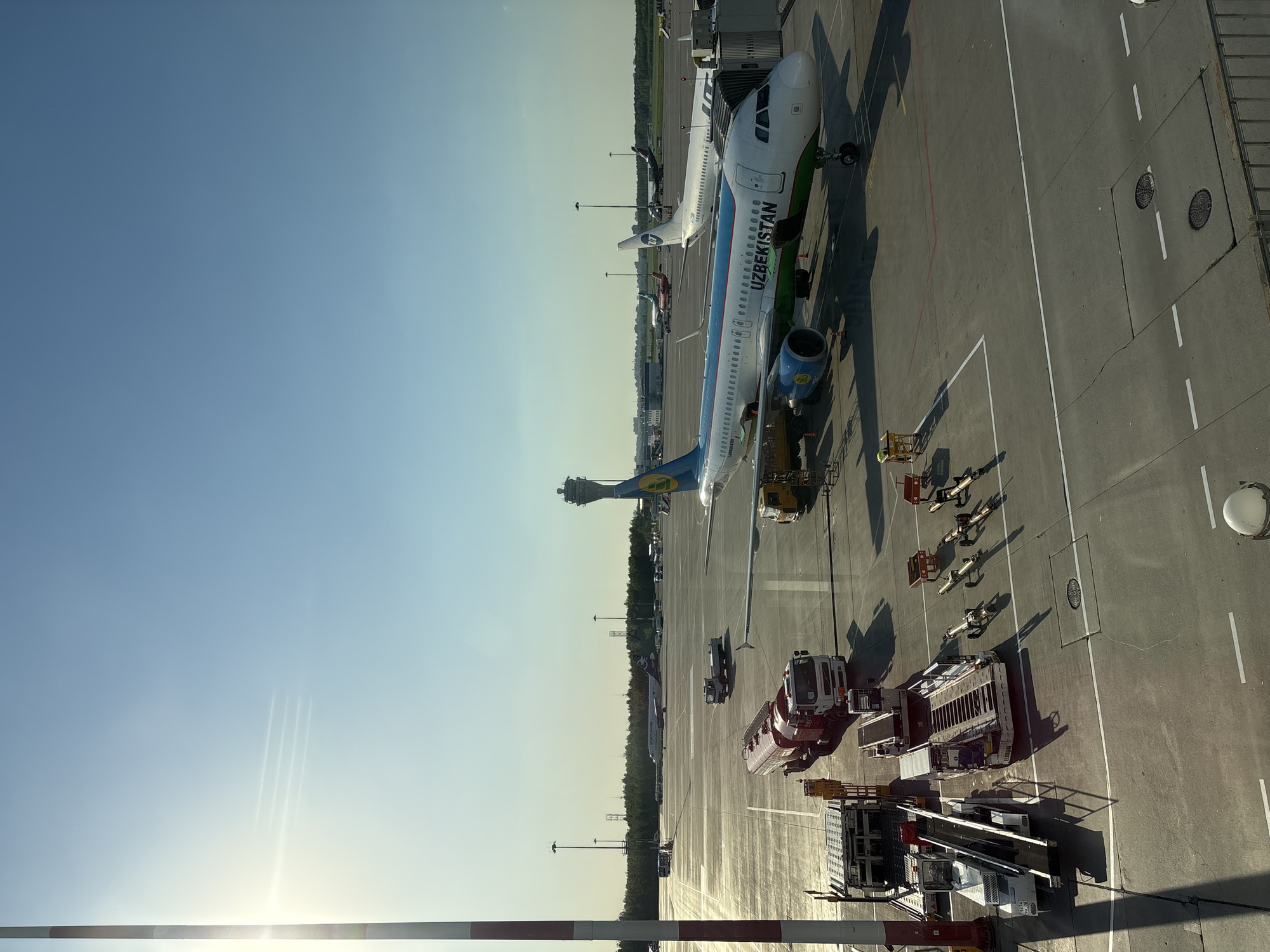 The image shows an aerial view of an airport tarmac. Two "Uzbekistan" branded passenger airplanes are parked next to each other at the terminal. Various ground support vehicles are nearby, including baggage handling equipment and refueling trucks. A fence is visible in the foreground, along with other airport structures. In the background, there are trees and the airport control tower. The sky is clear and blue. The location is Pulkovo International Airport in Saint Petersburg, Russia, as evidenced by the airplane markings and airport structures.
