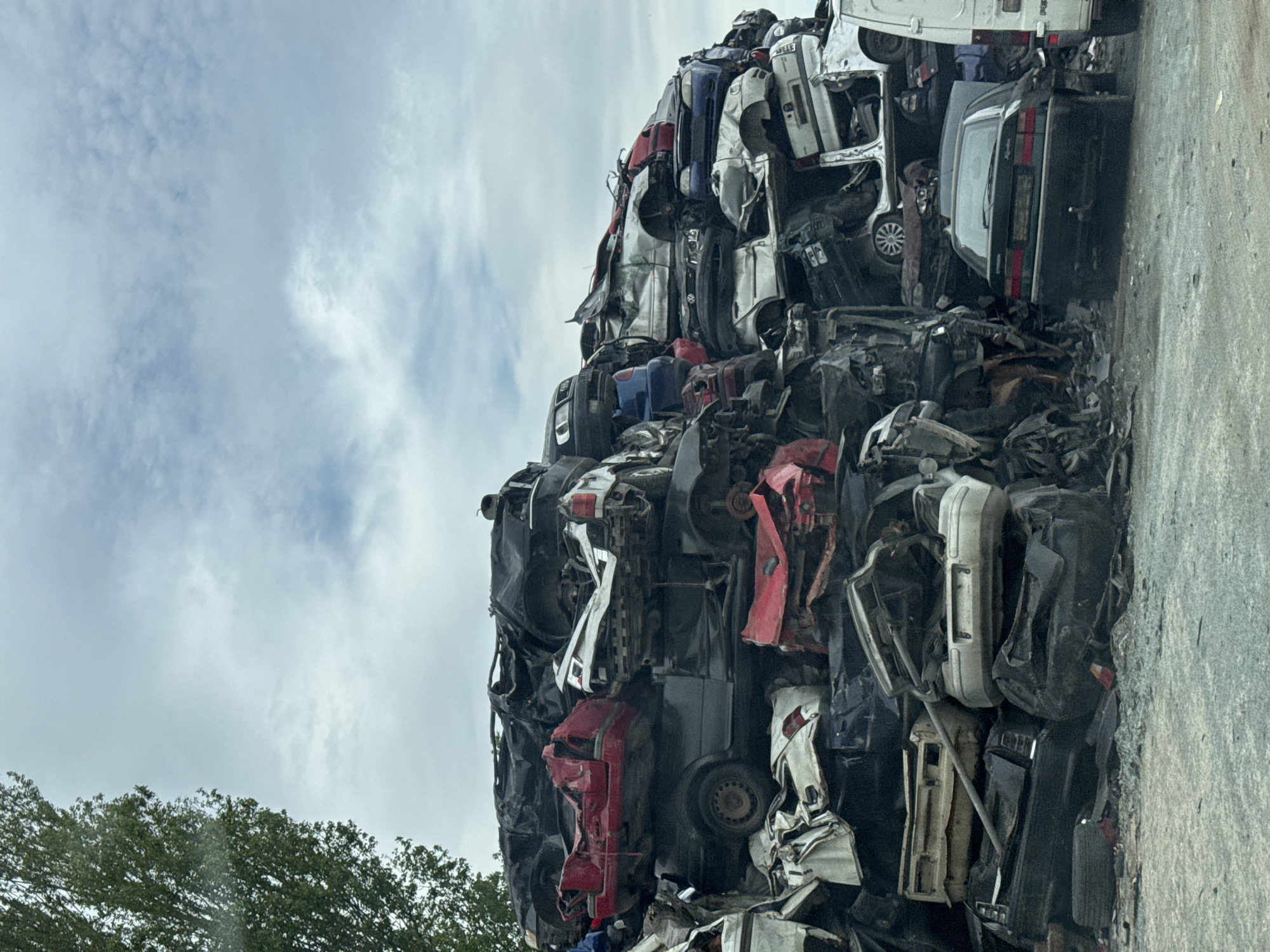 A vertical shot displays a large pile of crushed and stacked automobiles in a scrapyard. The vehicles are of varying colors - black, white, red, blue - and appear to be severely damaged and stripped. The pile is massive, reaching high into the sky where a cloudy sky is visible. A row of trees is visible in the bottom left corner of the image. The setting is a scrap yard, likely an industrial area in Varna, Bulgaria. There are no people visible in the image.