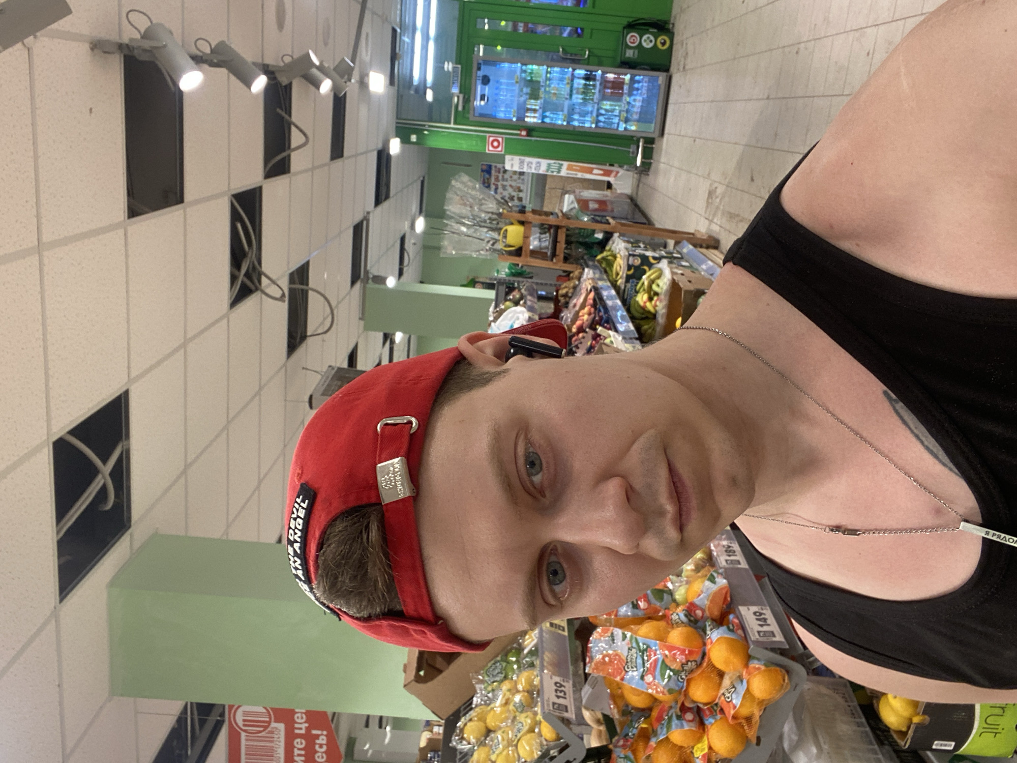 A young adult male is taking a selfie in what appears to be a grocery store in Samara, Russia. He is wearing a red baseball cap with a black patch that says "THE DEVIL AN ANGEL" and a black tank top. He has a silver necklace with a rectangular pendant. In the background, there are shelves stocked with fruits, including oranges and lemons, and a refrigerated display case with drinks. The ceiling has exposed wiring and fluorescent lighting. A green pillar is visible to the left of the man. The overall environment suggests a retail setting, likely a supermarket, within the city of Samara.