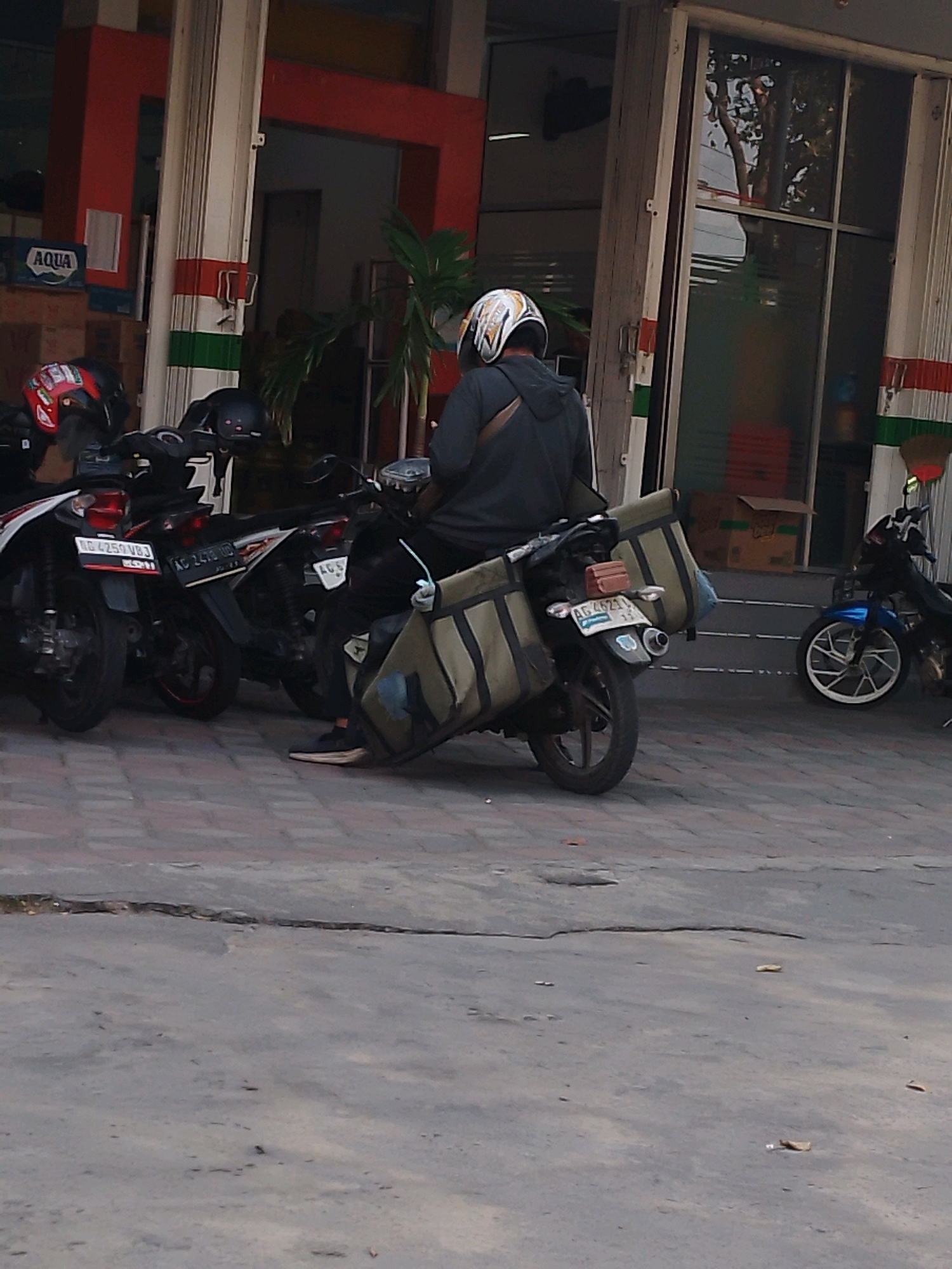 In Kertosono, Indonesia, a person wearing a gray hoodie and a helmet with white and black markings is sitting on a motorcycle with a large olive-green cargo bag attached to the rear. The motorcycle has a license plate reading "AG 4621 UD". The person is wearing dark pants and sneakers. The motorcycle is parked on a paved area with a crack in the foreground. In the background, there are other motorcycles parked, including one with a red and black helmet on it and another blue motorcycle with a distinctive white wheel. Adjacent to the motorcycles is a building with red and white trim and a sign that says "AQUA". The building appears to be a shop or business. The scene suggests a street or parking area outside a commercial establishment.