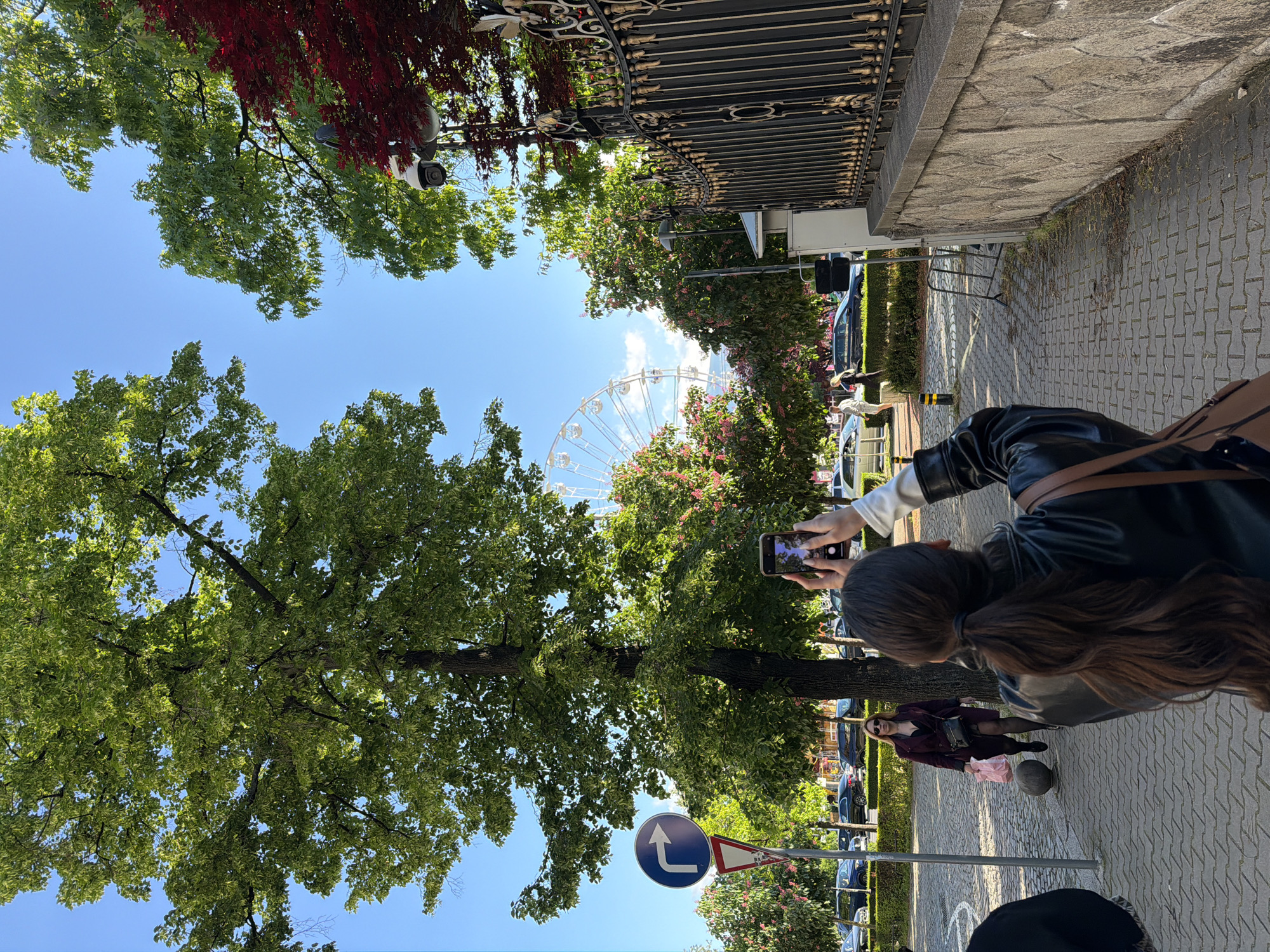 The image depicts a street scene in Sofia, Bulgaria, with a partial view of a Ferris wheel in the background, partially obscured by trees. A woman is capturing the Ferris wheel with her smartphone. In the foreground, a pedestrian walks along the pavement. A road sign indicating a left turn is visible. Cars are parked on the side of the street. Overhead, a security camera is mounted near a wrought-iron fence. Red foliage is visible near the fence. The sky is blue with some scattered clouds.