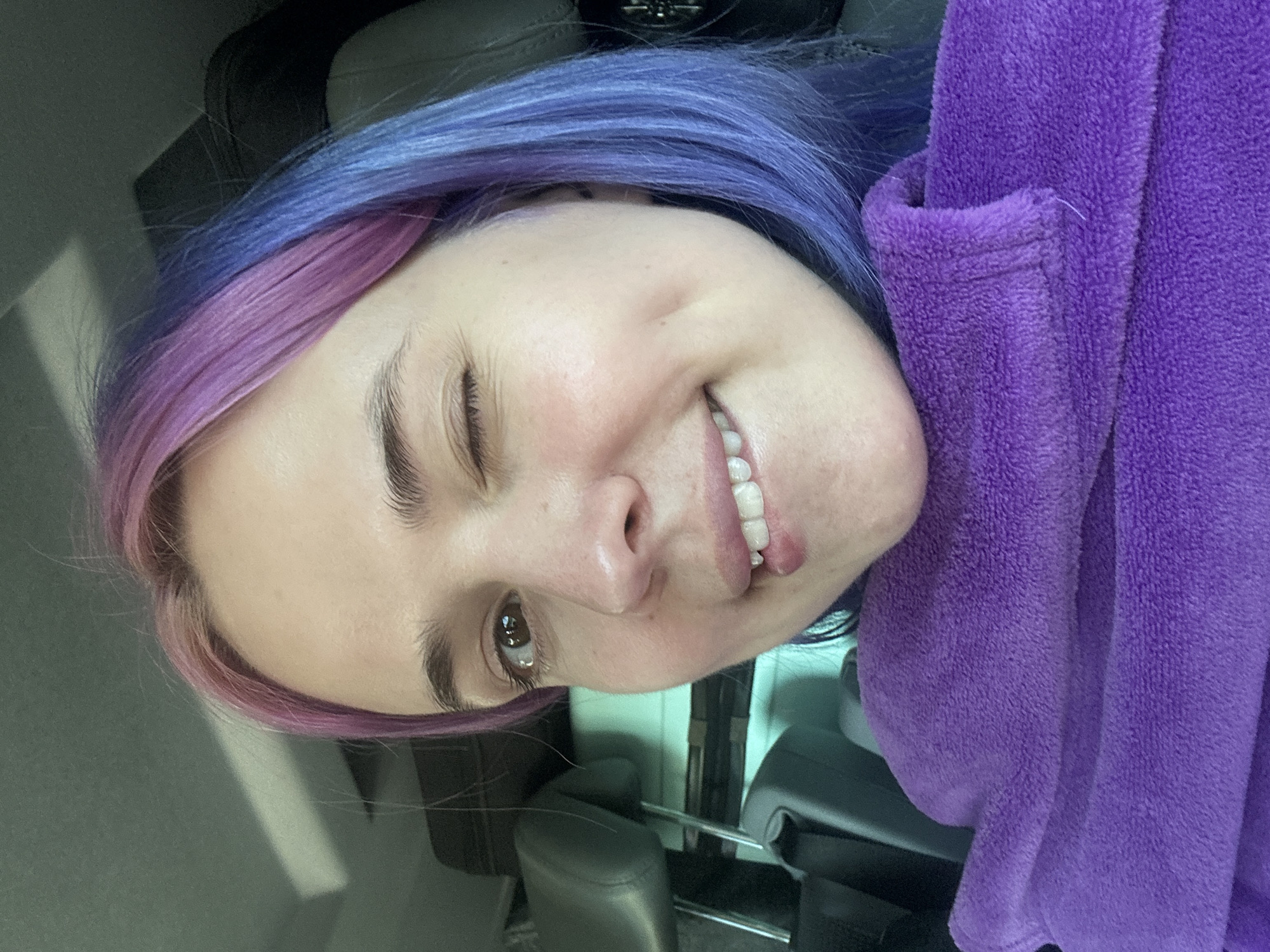 The image is a close-up, slightly angled selfie of a smiling woman with heterochromatic hair, colored purple and blue, who is winking her right eye. She is wearing a purple, textured garment. The setting appears to be inside a car based on the visible headrest and interior features. The location within the city of Le Muy, France, cannot be determined from the visual data. No specific actions or interactions can be discerned beyond the subject taking a selfie.