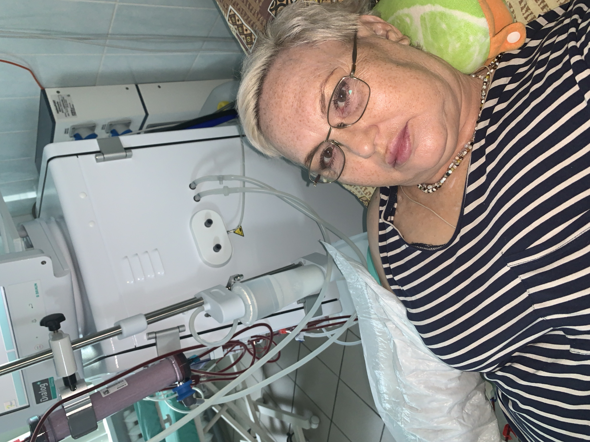 The image depicts an older woman lying down next to a dialysis machine. She is wearing glasses and a striped shirt, with a pillow behind her head. The dialysis machine is white and has various tubes and components visible. The setting appears to be a home or medical facility. The location is presumed to be Chaikovsky, Russia.