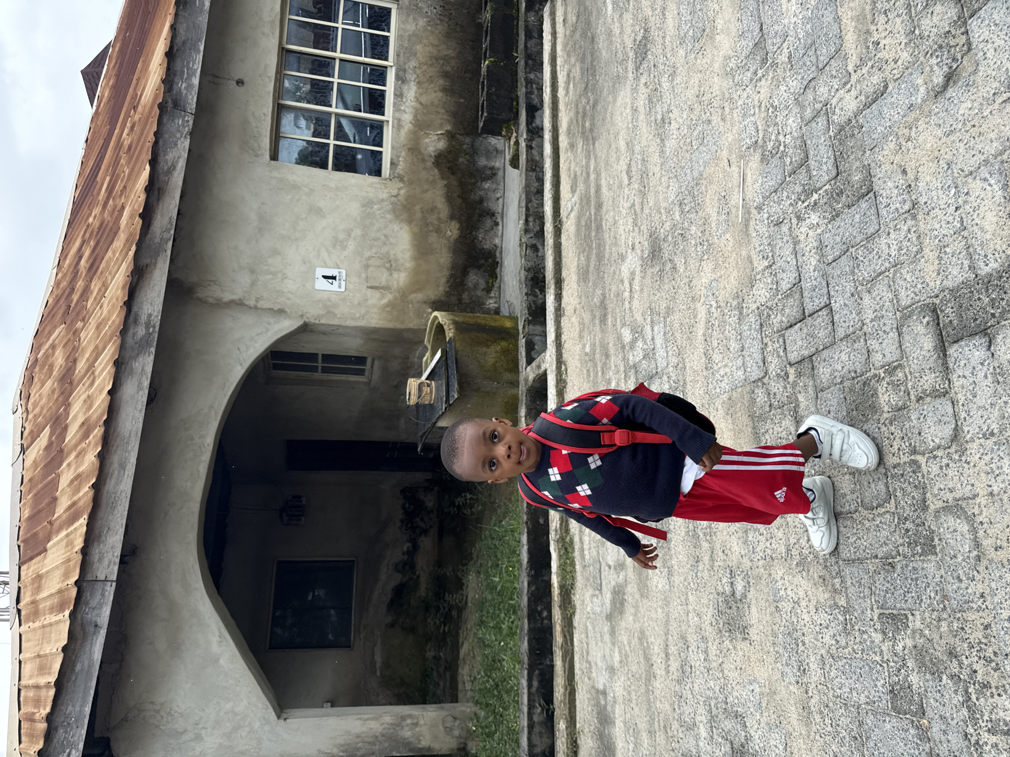 In Ado, Nigeria, a young boy with dark skin is standing on a paved courtyard in front of a weathered building. The boy is wearing a dark blue sweater with a diamond pattern in red, green, and white, red shorts with white stripes down the sides and the Adidas logo, and white Velcro sneakers. He has a red backpack on. The building is constructed of rough, gray concrete and features a corrugated metal roof with areas of rust. To the left of the boy, there is an arched entryway leading into the building, and a window is visible within the arch. To the right of the entryway, a multi-paned window is present. Adjacent to the building, there is a round concrete well or cistern, and beyond that, several dark concrete blocks are arranged. The ground is paved with rectangular gray stones laid in a herringbone pattern. The sky above is overcast.
