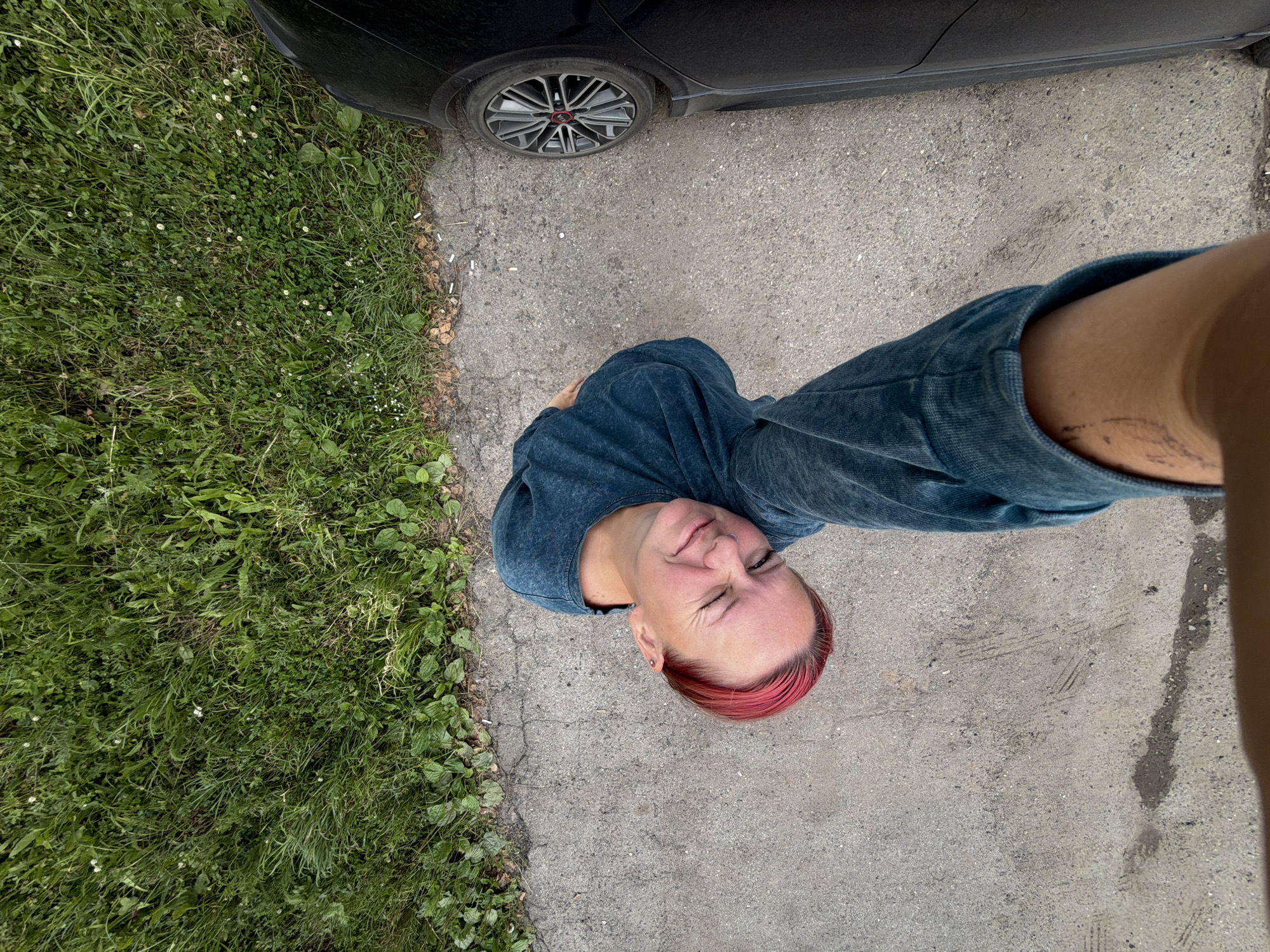 This is a candid, high-angle, full-body shot of a person standing on a paved area next to a patch of grass. The person is wearing a dark blue top and blue jeans. They have short hair dyed pink at the ends and are winking. A black car is partially visible in the top left of the frame, with its front wheel and fender. The ground is a mix of concrete and grass, with some small white flowers scattered in the grass. There are also a few cigarette butts on the edge of the pavement. The lighting suggests it is daytime, possibly overcast, given the soft shadows. The location cues, such as the car and the type of pavement and vegetation, are consistent with a suburban or urban environment in Izhevsk, Russia. The person's expression appears playful or mischievous. There is no visible text in the image.