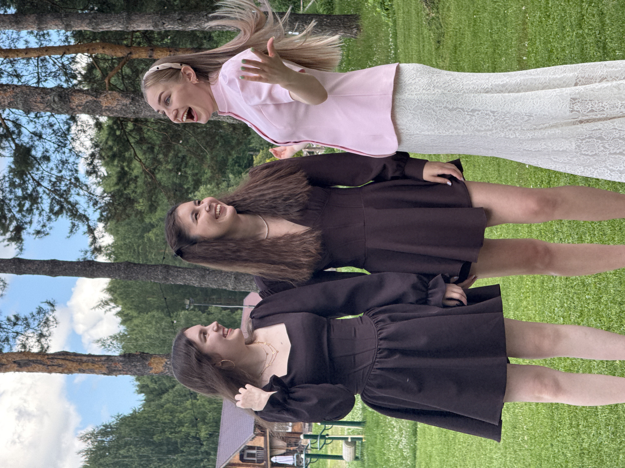 Three young women are standing in a grassy outdoor area, possibly a park or a backyard, in Votkinsk, Russia. The woman on the left is wearing a dark brown long-sleeved dress with a square neckline and a flared skirt. She is looking to her left and smiling. The woman in the middle is wearing a similar dark brown dress, also with a flared skirt, and is looking to her right and smiling. The woman on the right is wearing a pink collared shirt with short sleeves and a white lace skirt. She is looking upwards and laughing, with her hands raised. Behind them, there is a wooden building with a brown roof, some trees, and a cloudy sky. The ground is covered in green grass. The scene appears to be a candid moment of friendship and enjoyment.