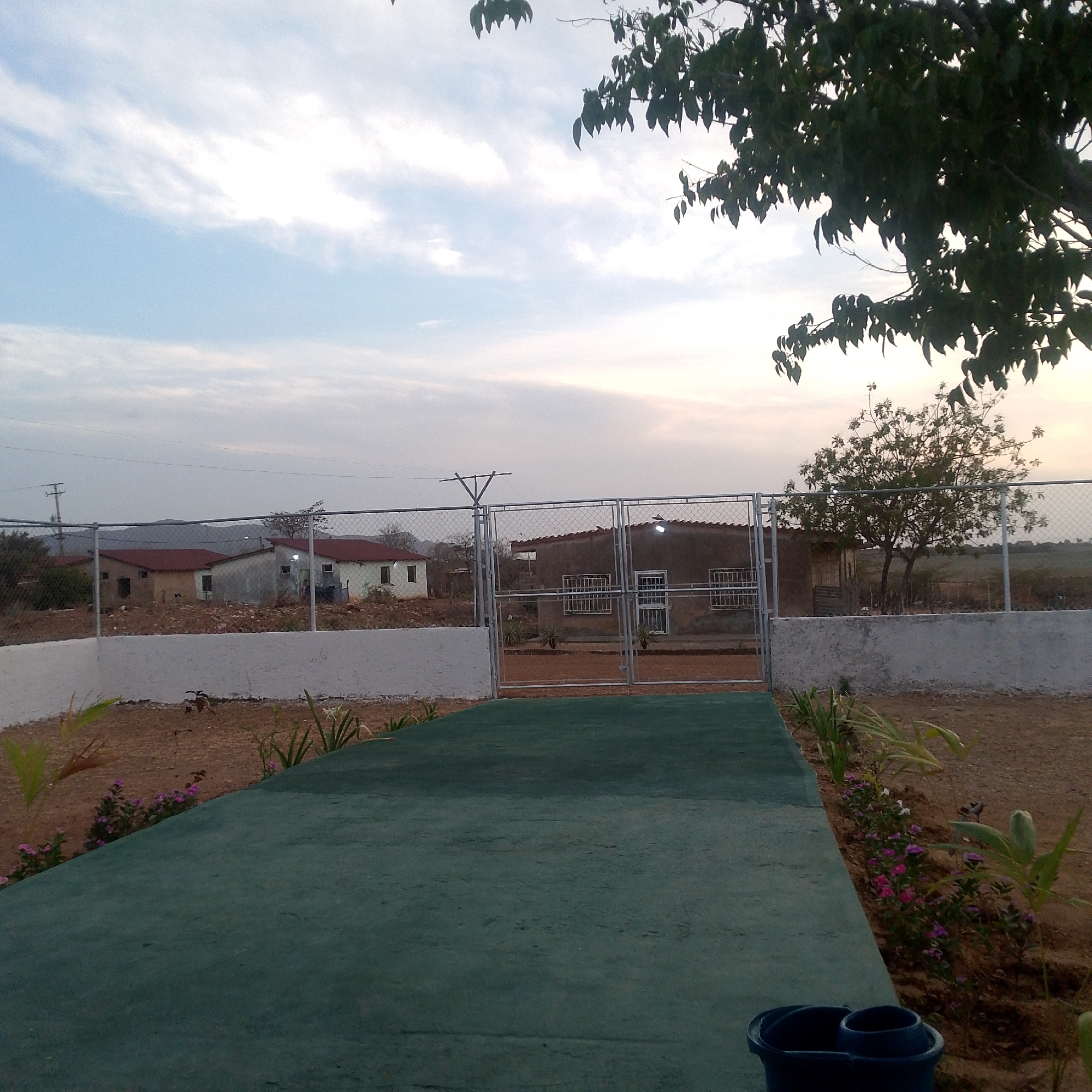 The image shows a partially obscured view of what appears to be a park or garden setting in Altagracia, Venezuela. In the foreground, there is a green concrete path leading towards a gated fence. The fence encloses a structure, possibly a building or house, with windows and a door visible. On either side of the path, there are planted flowers and other greenery. Beyond the fence, there are additional buildings, trees, and in the distance, what appear to be mountains. The sky is overcast, suggesting early morning or late afternoon lighting conditions. A blue container is visible in the lower right corner. The overall scene gives the impression of a tranquil, semi-rural environment.