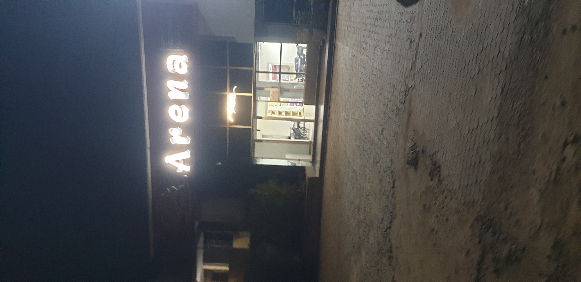 The image shows the exterior of a building at night, with the word "Arena" lit up in neon letters above an entrance. The entrance appears to be a glass-fronted doorway leading into a well-lit interior. Inside, tables and chairs are visible, suggesting the space might be a restaurant or a recreational area. The ground in front of the building is paved with interlocking bricks, and there is a patch of what appears to be dirt or soil on the left side of the image. The lighting suggests it is dark outside, with the neon sign and interior lights being the primary sources of illumination. No people are clearly visible in the image. The location in Narayi, Nigeria, is not explicitly indicated by any visible landmarks or text in the image itself, but is provided as contextual information.