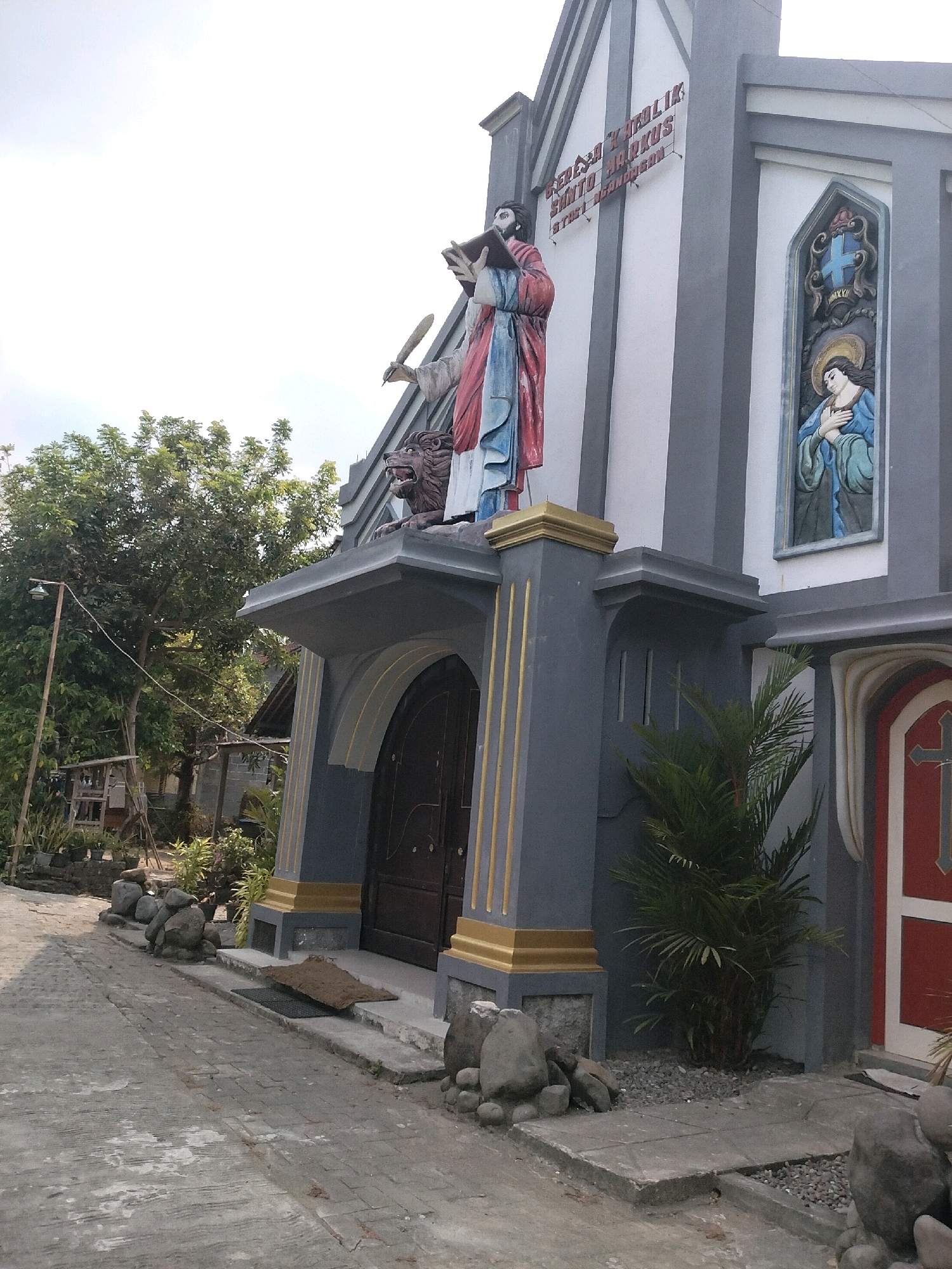 The image shows the exterior of the St. Mark Catholic Church in Ngandangan, Indonesia. The building has a gray facade with white accents and is adorned with religious statues and artwork. A statue of a robed figure holding a book and quill stands prominently above the entrance. To the right, a stained-glass window depicts a religious scene. The church is surrounded by greenery and a paved walkway. The scene appears to be a sunny day, with a bright sky overhead.